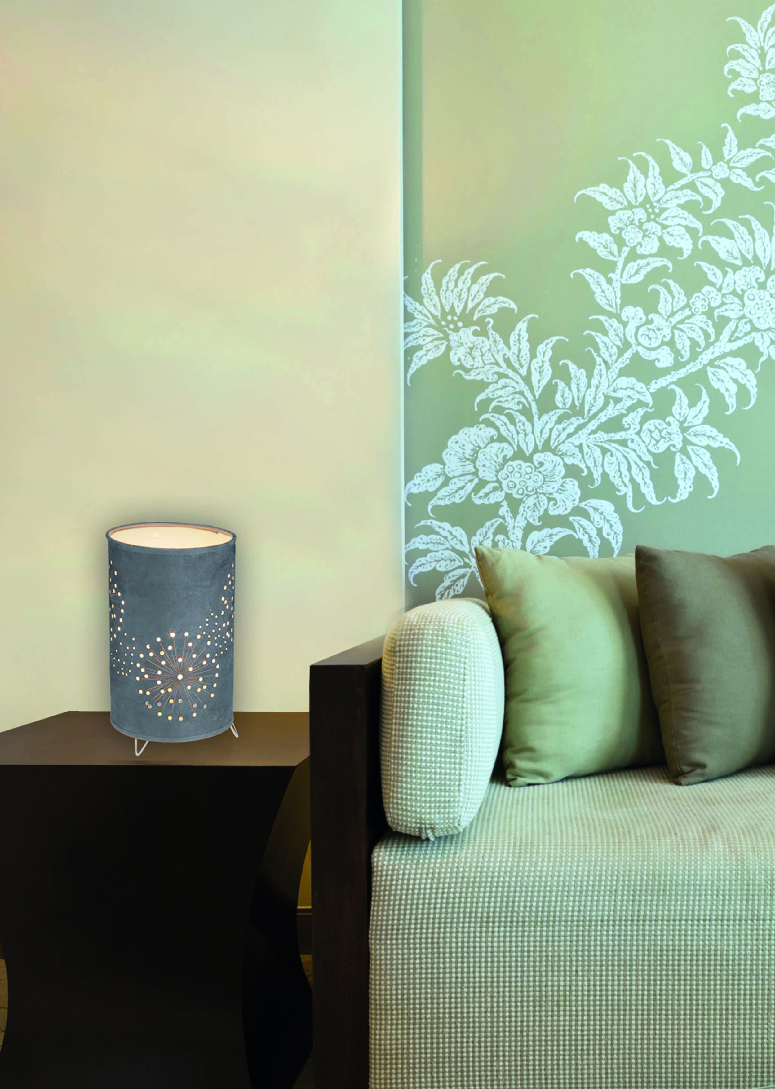 Graue Lampe mit Blumendesign auf einem Tisch neben einem Sofa mit Kissen und Wand.