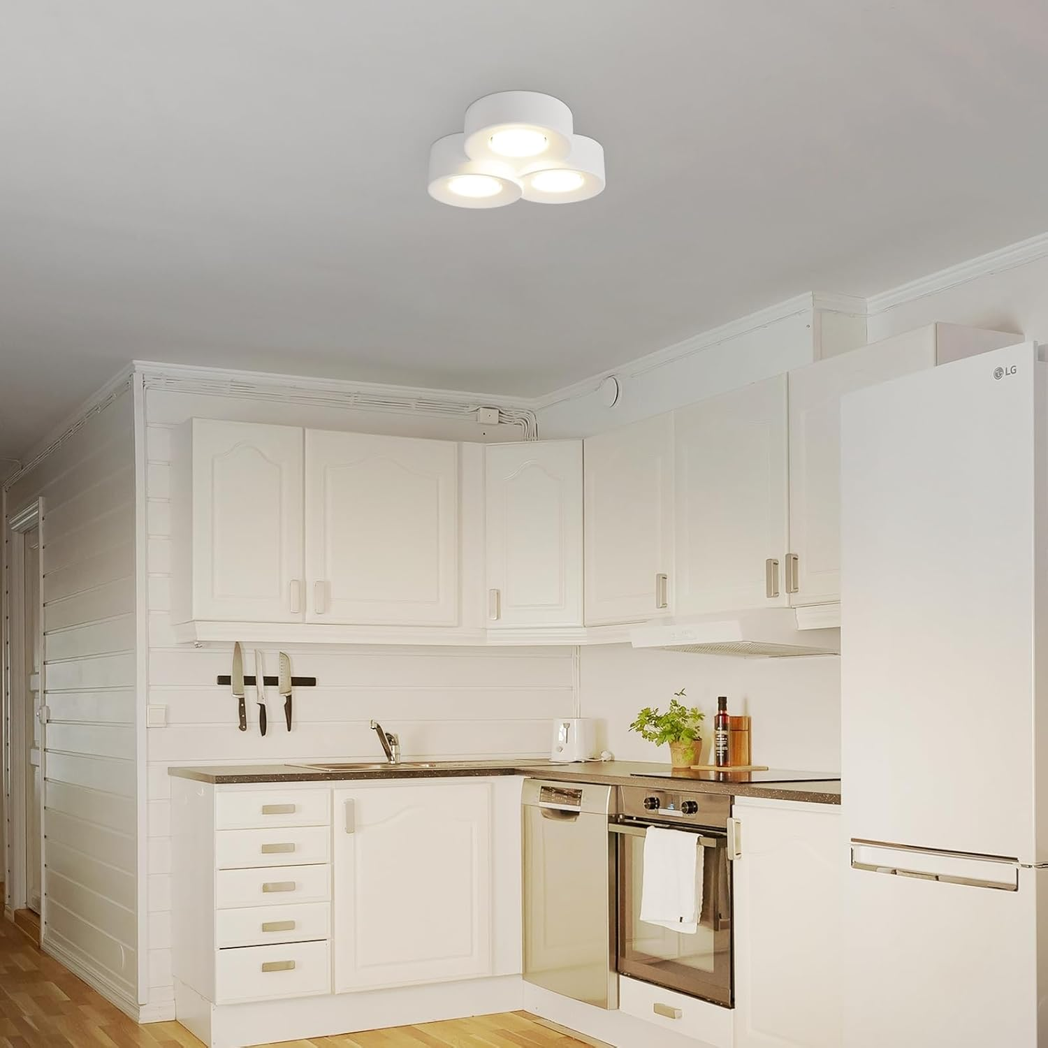 Una cocina blanca con luces de techo. Armarios blancos y un fregadero y electrodomésticos de acero inoxidable.