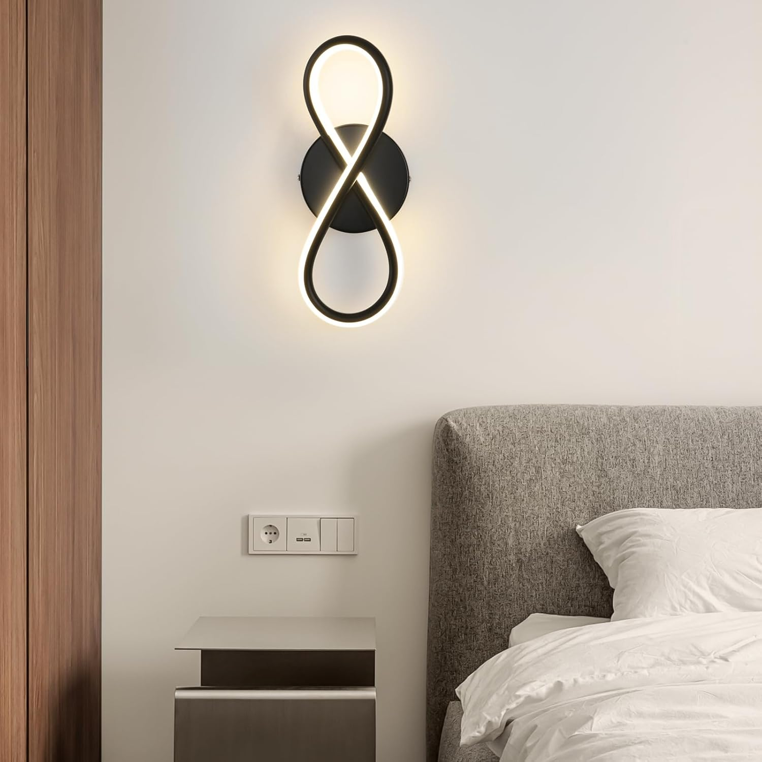 Una moderna lampada da parete nera a forma di simbolo dell'infinito, montata su una parete bianca sopra un comodino.