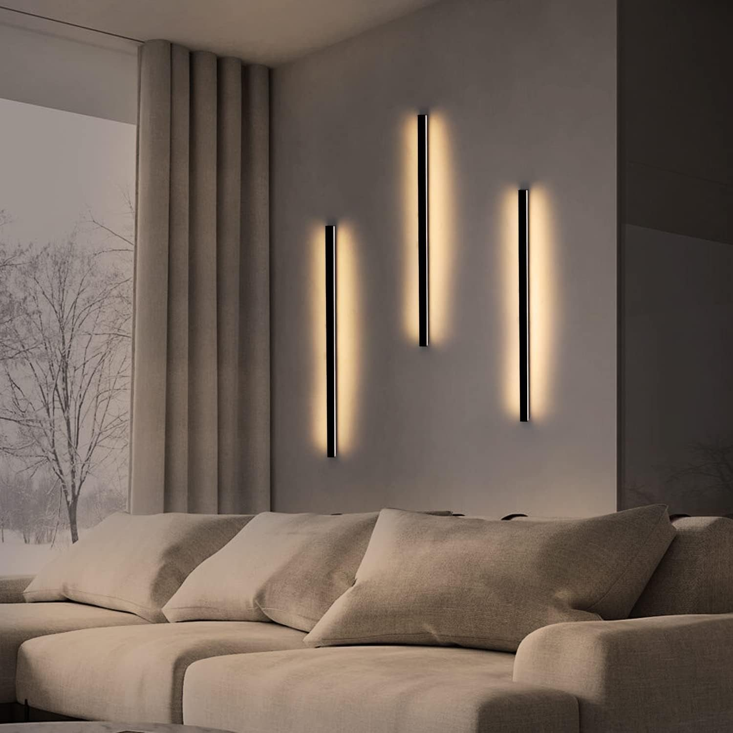 Tre lampade da parete nere verticali con luce calda, sopra un divano beige. Sfondo: parete, finestra, tenda.