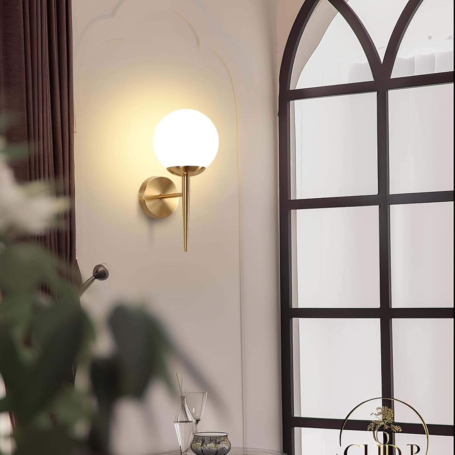 Una lámpara de pared dorada con una luz de globo blanca. Una ventana y una planta están en el fondo.