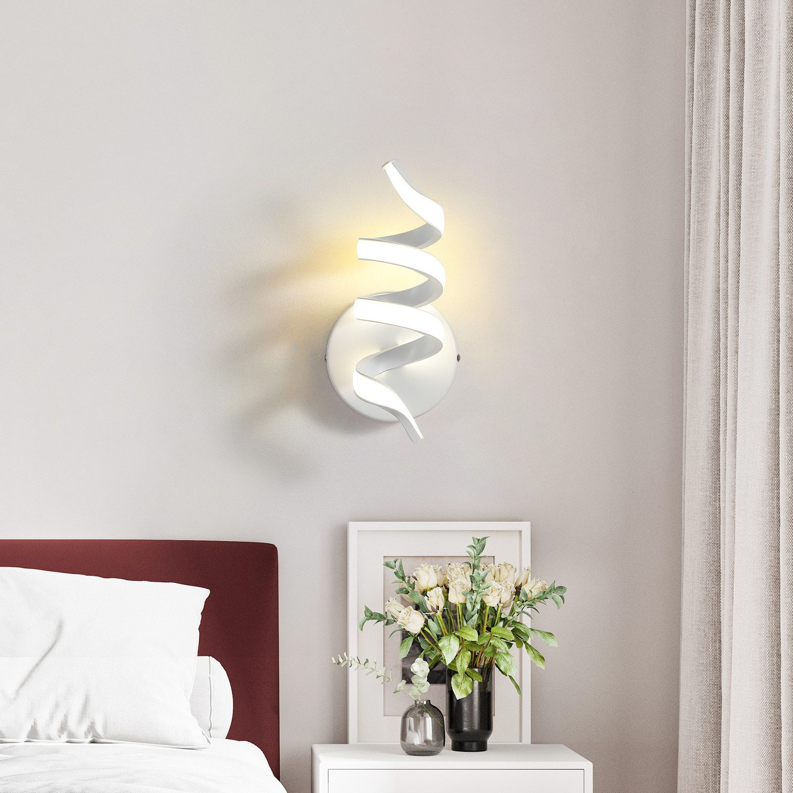 Una lampada da parete a spirale bianca illuminata, accanto a un comodino con fiori e un quadro.