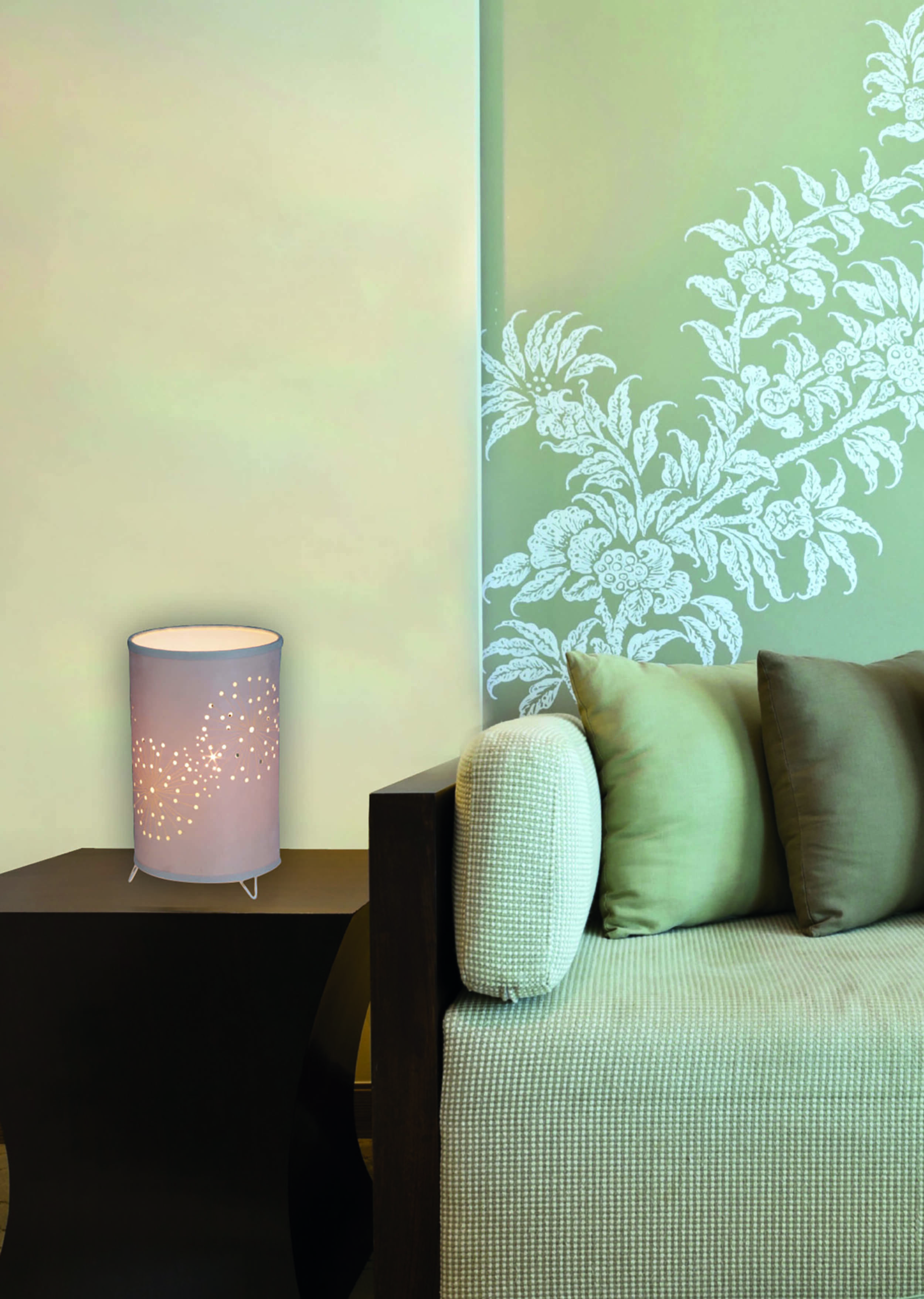 Eine zylindrische Lampe mit sternförmigen Aussparungen steht auf einem dunklen Tisch. Im Hintergrund ein Sofa mit Kissen.