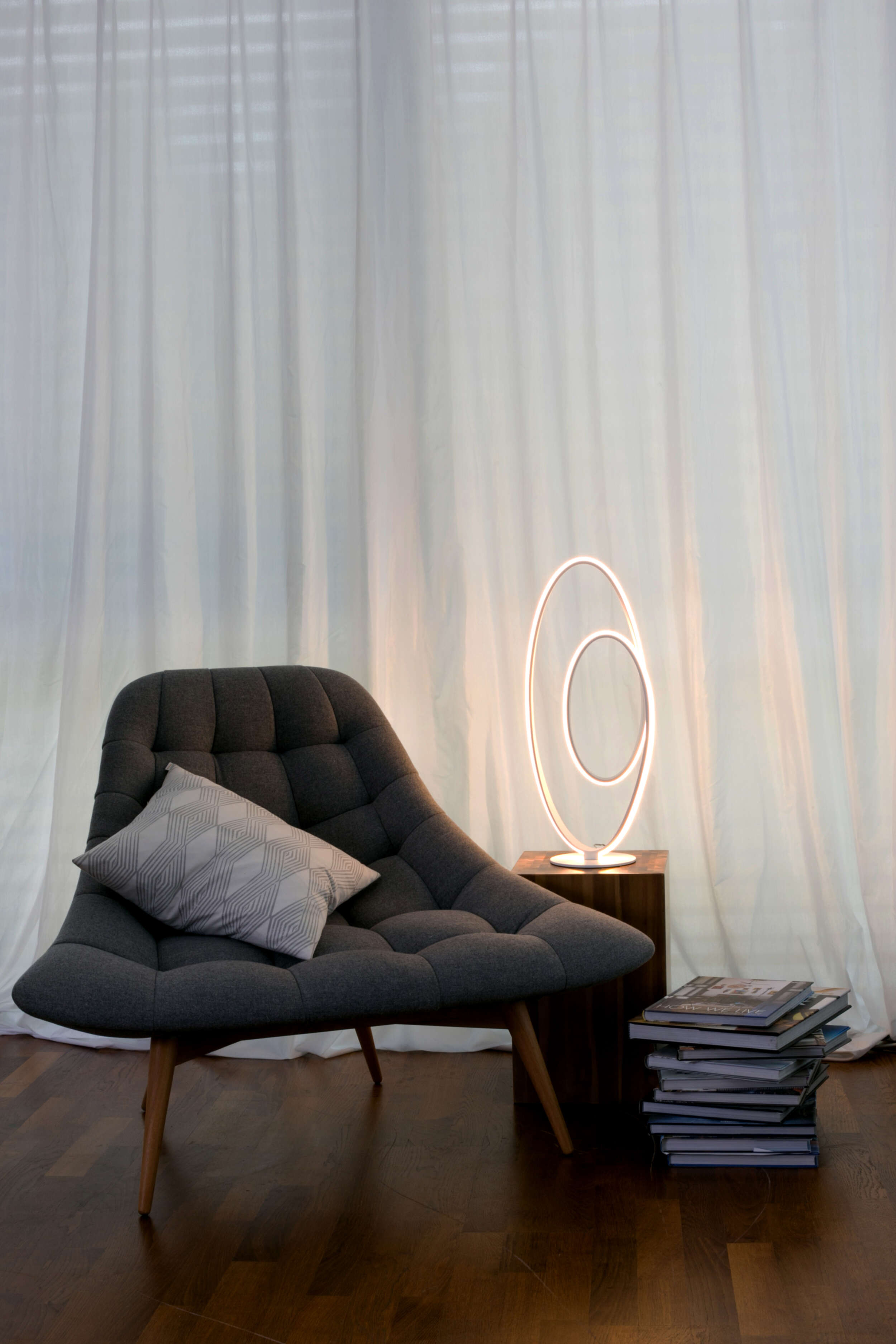 Moderne Lampe neben Stuhl und Bücherstapel, mit weißem Vorhang im Hintergrund.