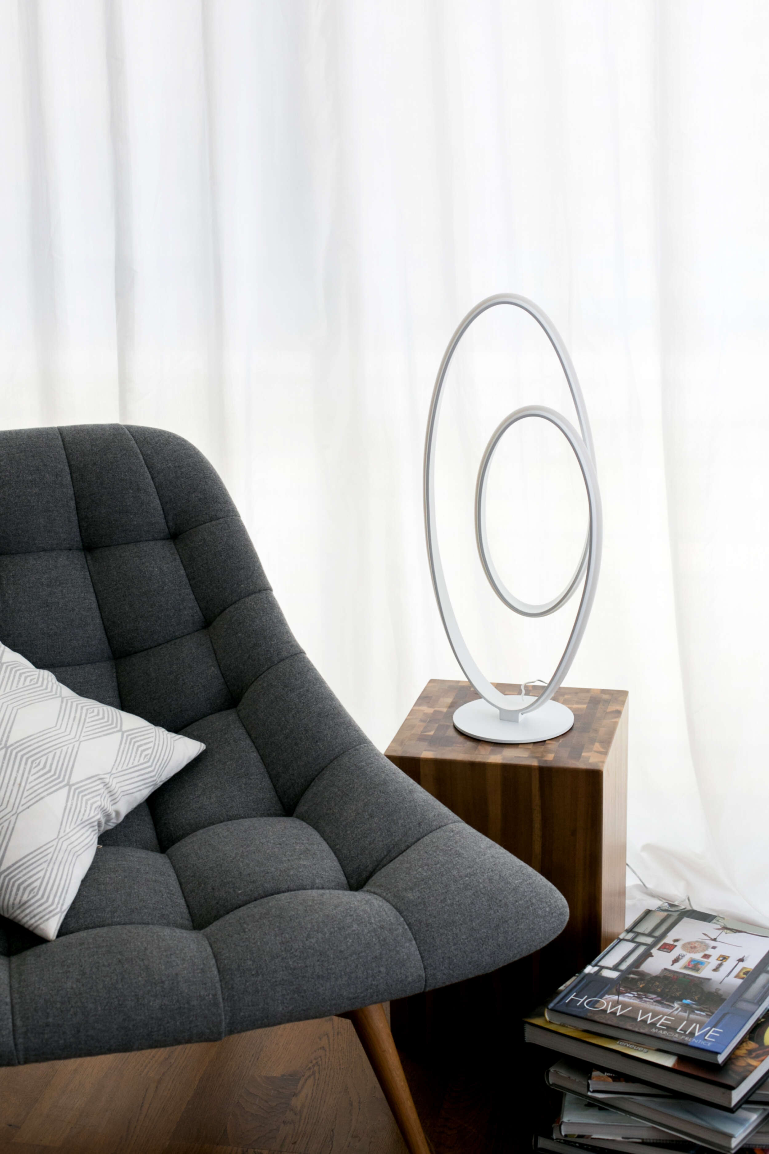 Eine moderne Lampe auf einem Holzblock, neben einem grauen Stuhl mit Kissen und einem Bücherstapel.