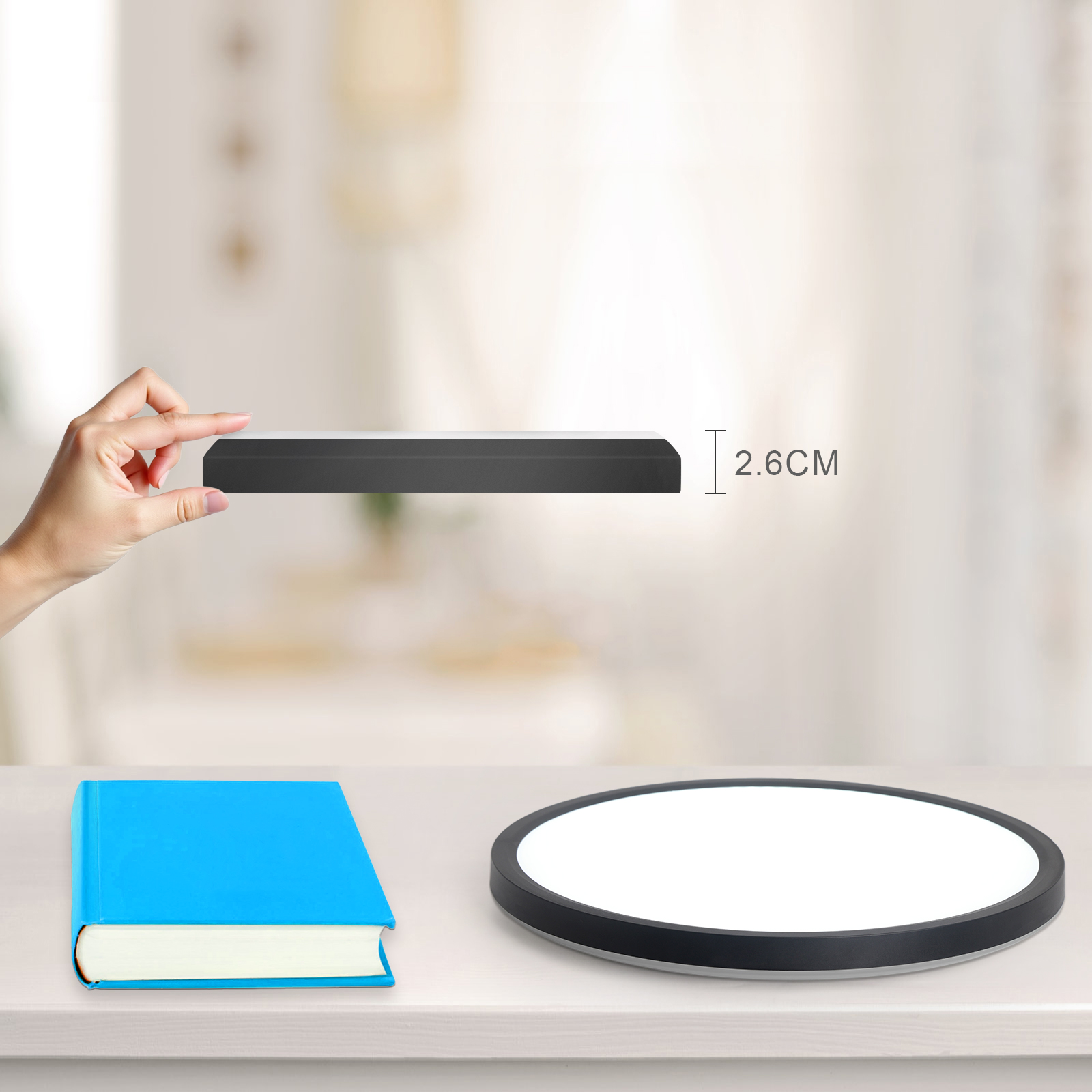 Mano sosteniendo un objeto delgado y una luz de techo redonda con un libro azul sobre una mesa.