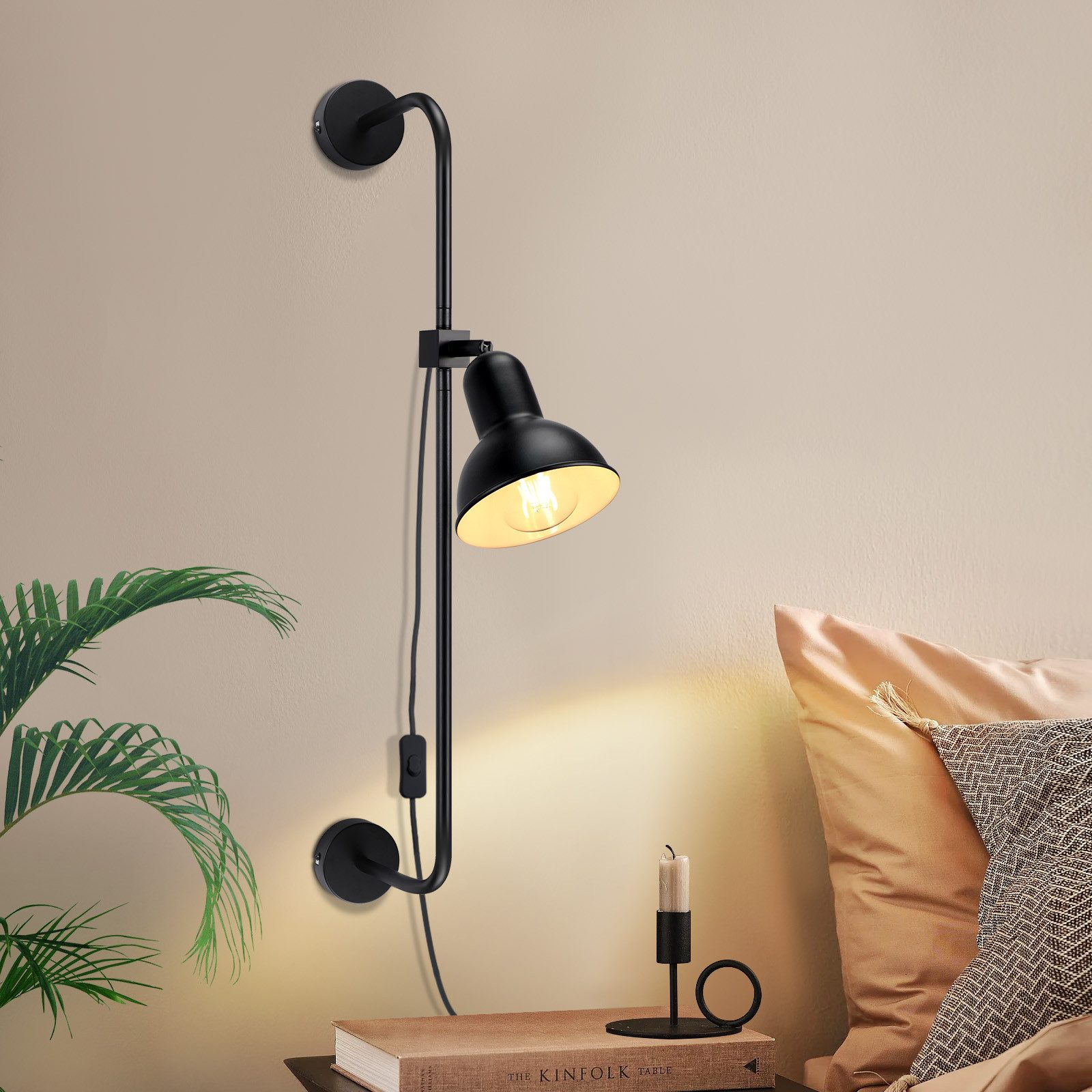 Lampada da parete nera con braccio regolabile, accanto a un libro e una candela. Muro bianco.