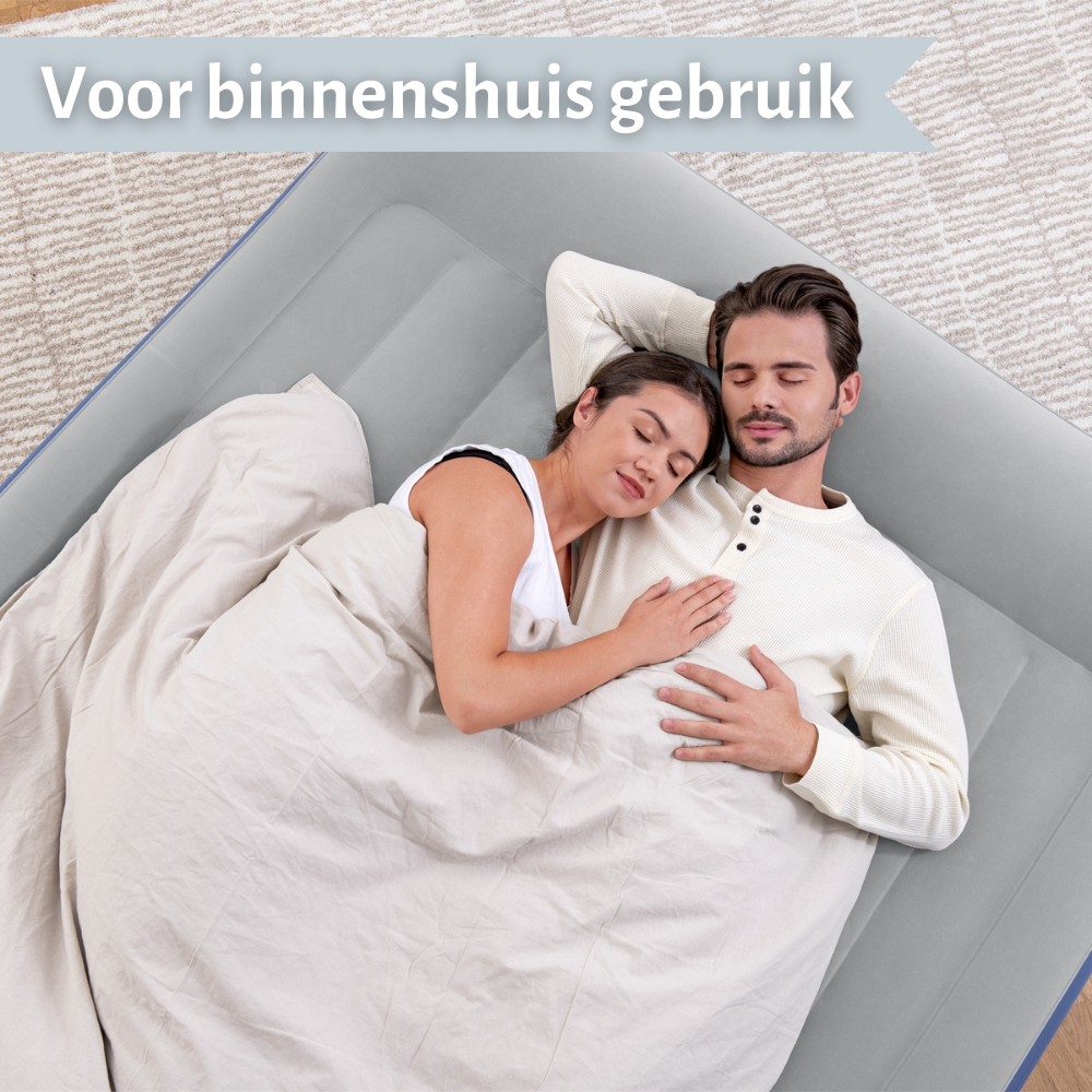 Een stel slaapt op een grijze luchtmatras, bedekt met een deken. De scène is binnenshuis, met een lichtgrijze achtergrond.