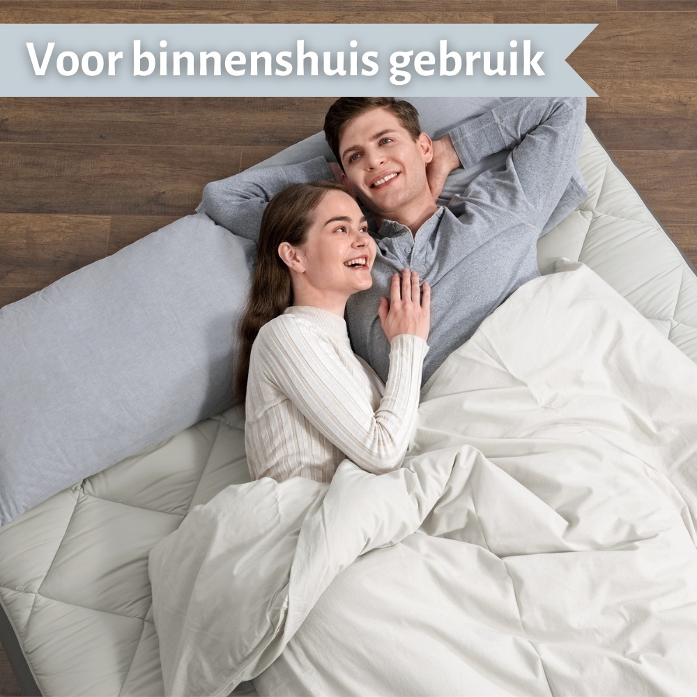 Un couple allongé sur un matelas pneumatique couvert d'une couverture, souriant. Le fond montre un sol et un mur en bois.