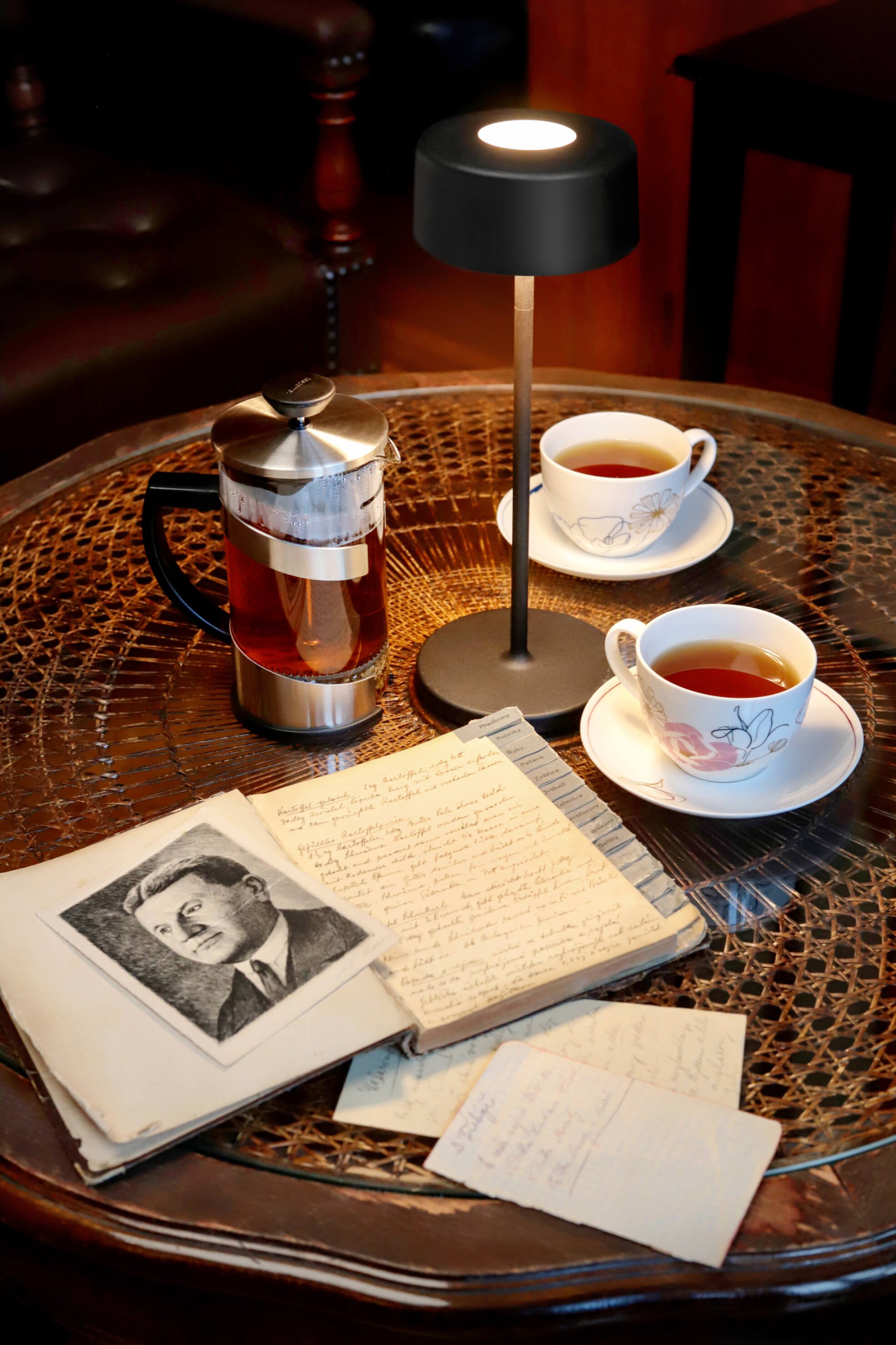 Tisch mit Lampe, Tee und offenem Buch. Eine French Press und Teetassen stehen auf dem Tisch.