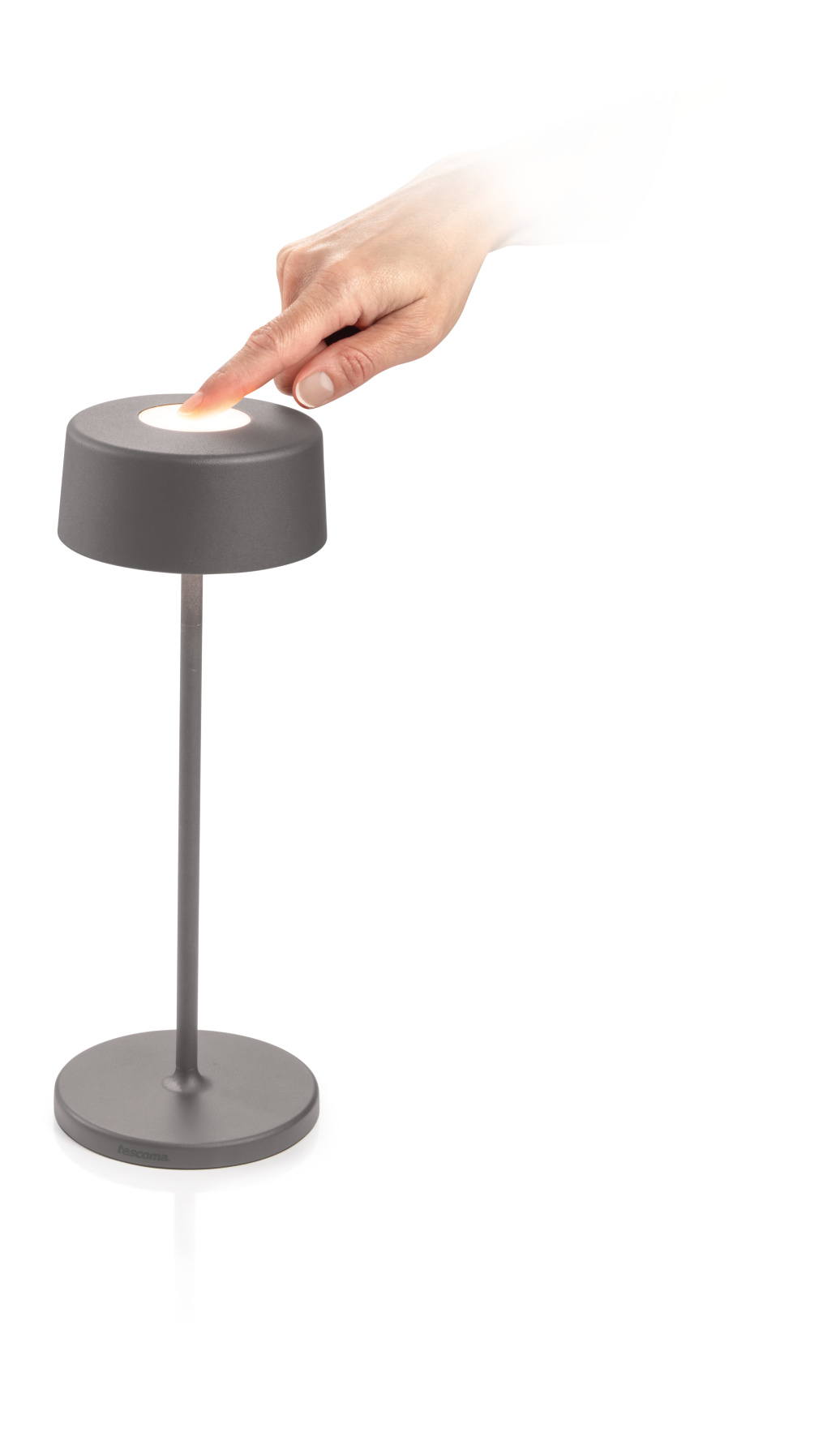 Eine Hand berührt eine graue Lampe mit rundem Fuß. Die Lampe ist an und strahlt sanft.
