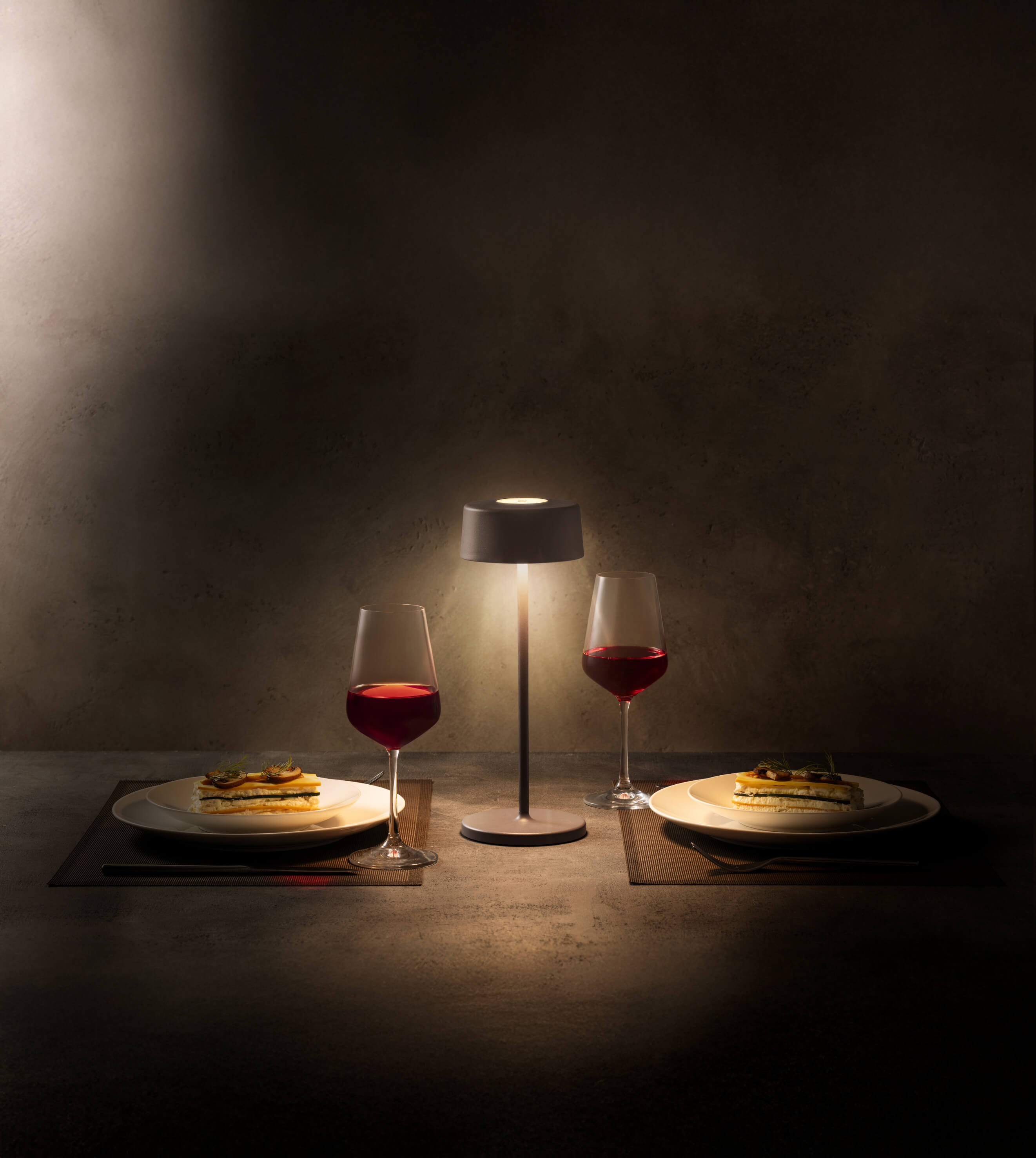 Romantisches Abendessen mit zwei Gläsern Rotwein, Tellern und einer kleinen Lampe auf einem Tisch.