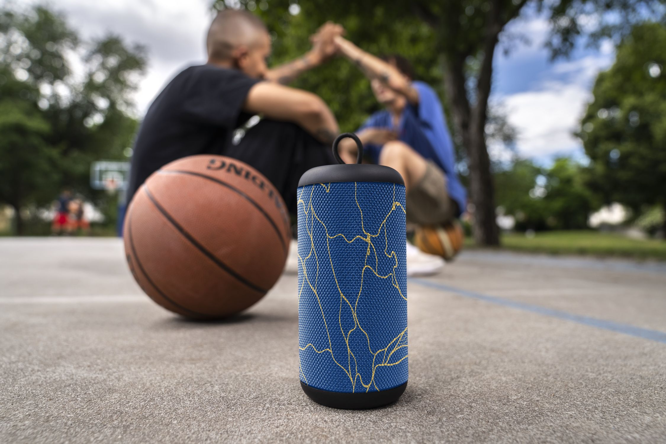 Głośnik w niebiesko-złote wzory na betonie z dwoma rozmytymi koszykarzami w tle.