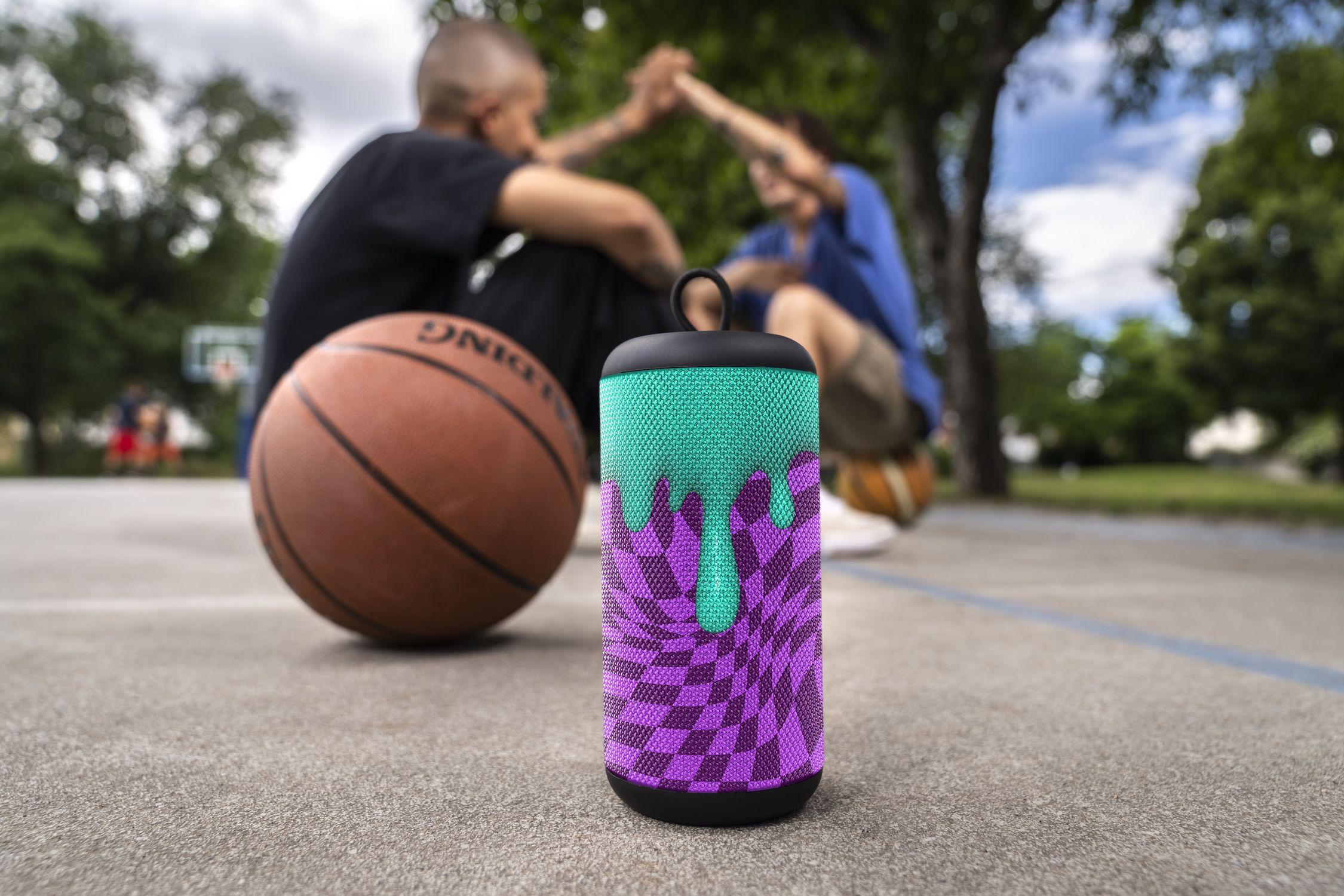 Głośnik z zielonym i fioletowym wzorem, przed dwiema osobami i koszykówką na boisku.