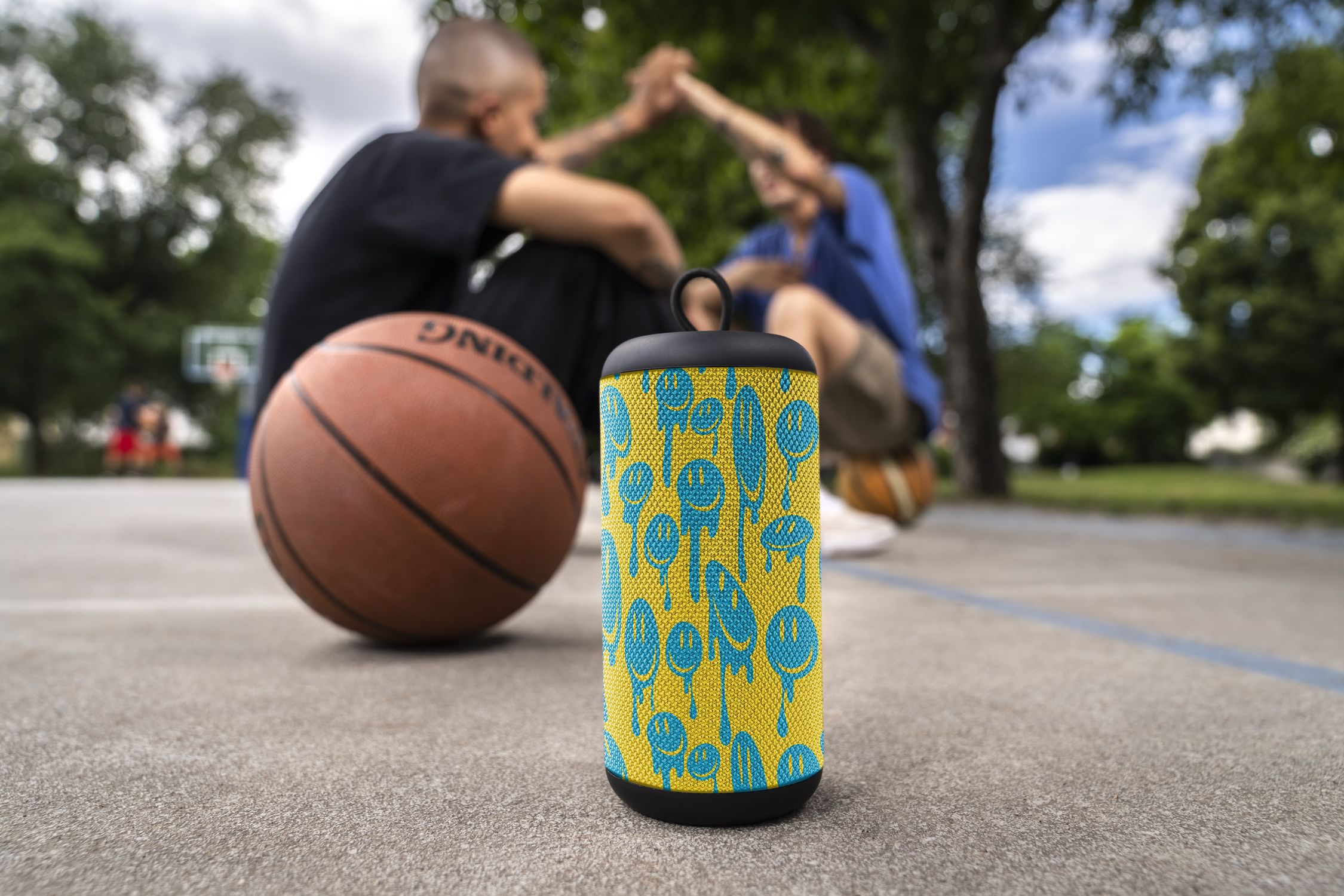 Głośnik z żółtym i niebieskim wzorem, przed dwiema osobami i koszykówką na boisku.