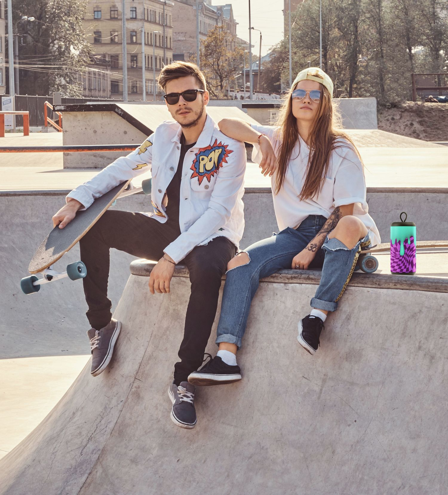 Dwie osoby w okularach przeciwsłonecznych i deskorolkami siedzące na rampie skateparku.