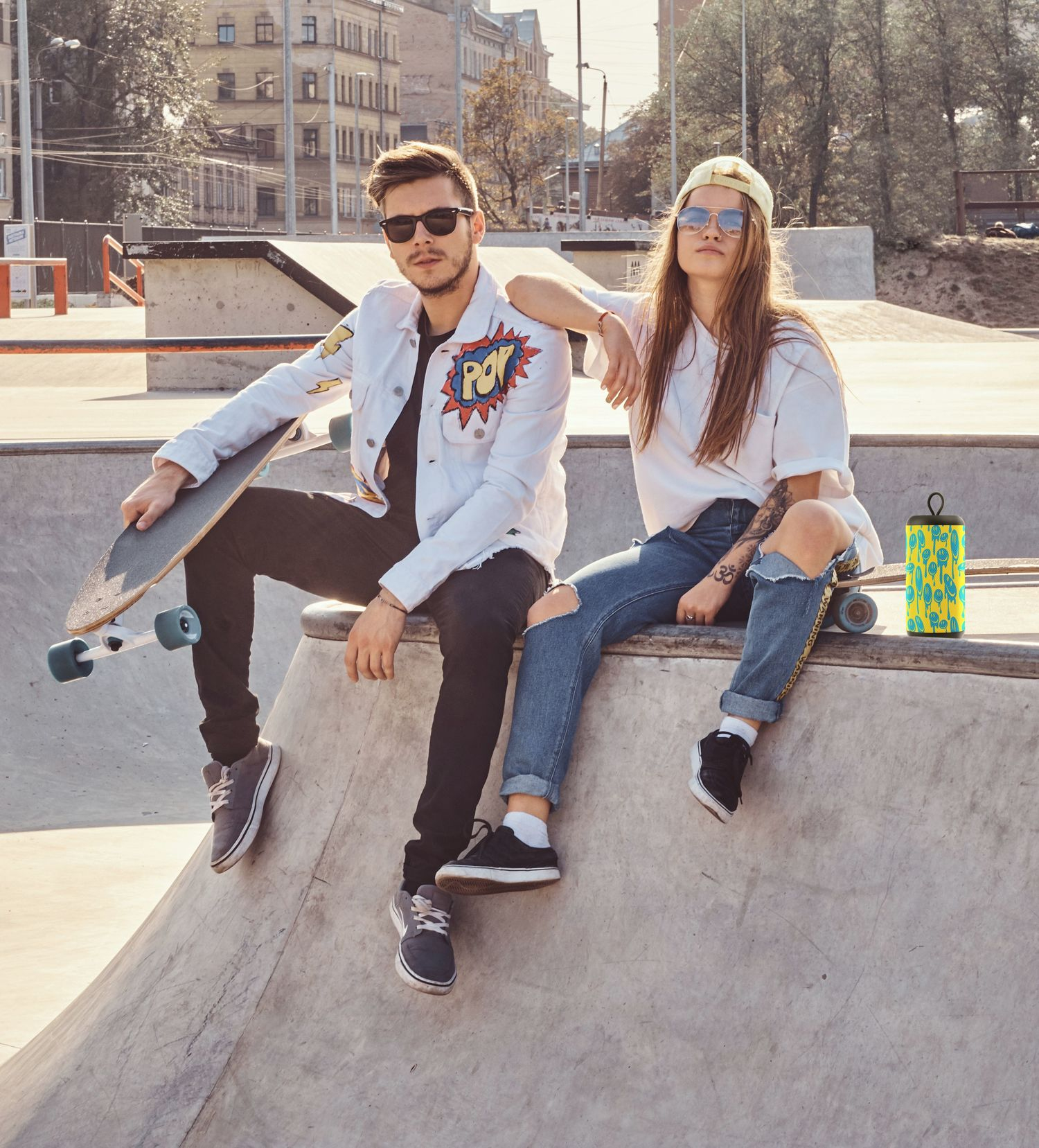 Dwie osoby siedzące na krawędzi skateparku z głośnikiem i deskorolką.