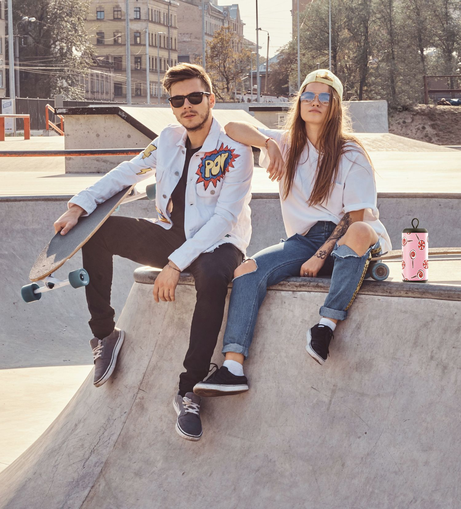 Dwie osoby siedzące na skateparku. Mężczyzna trzyma deskorolkę, a kobieta ma różowy kubek.