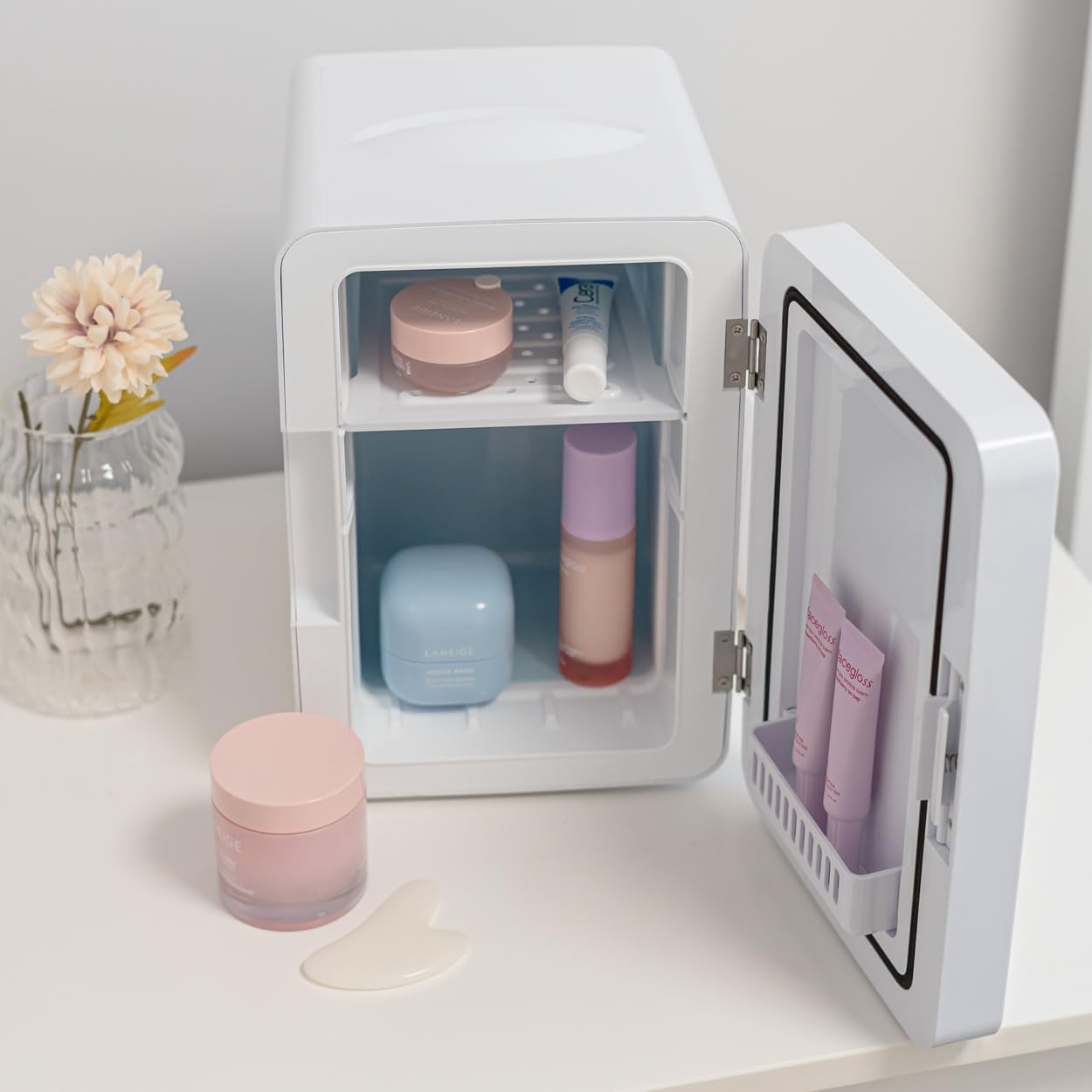 Un mini-refrigerador blanco con puerta abierta, contiene cosméticos. Un florero a la izquierda.