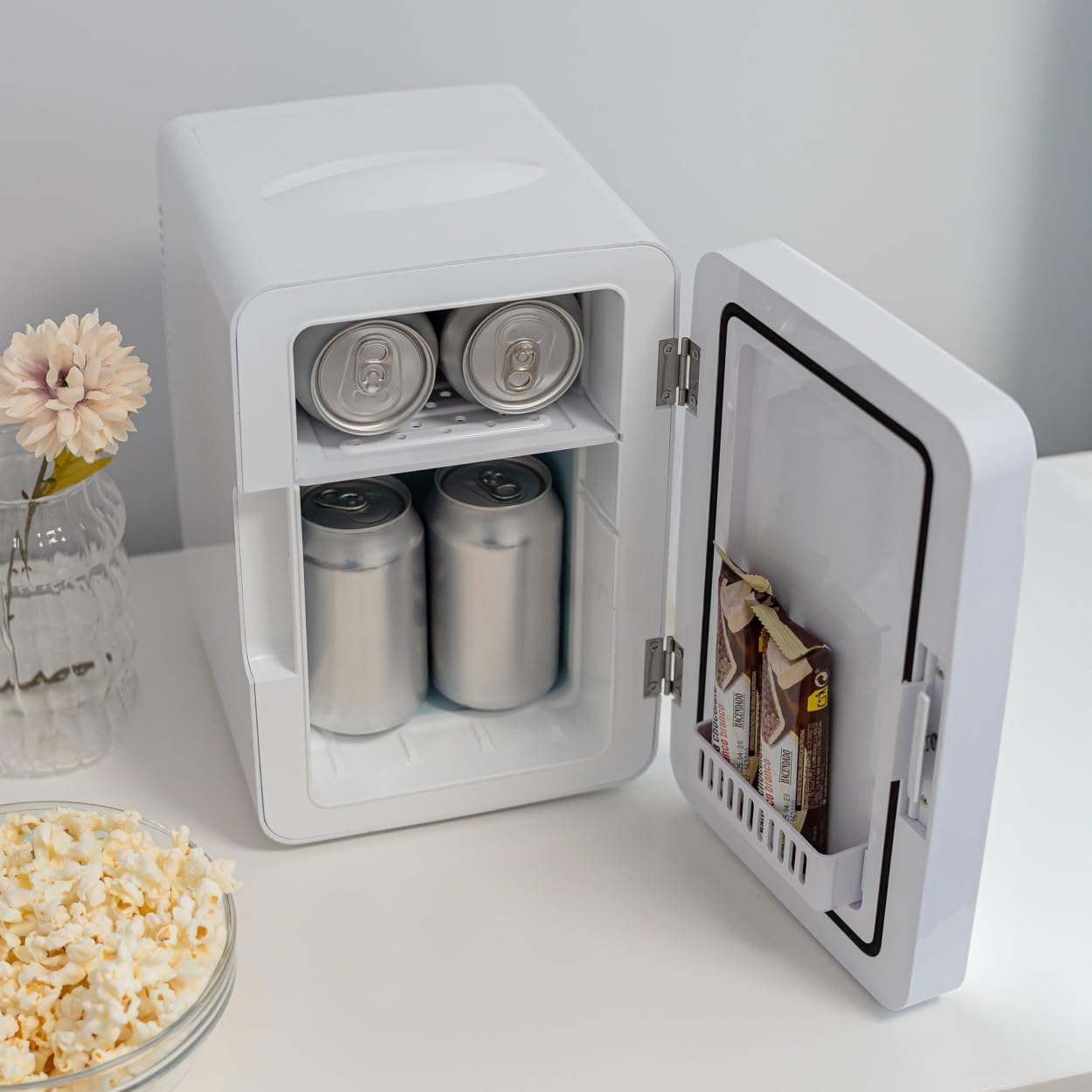 Mini-nevera blanca con puerta abierta, latas y snacks dentro. Palomitas y un jarrón con flores en la mesa.