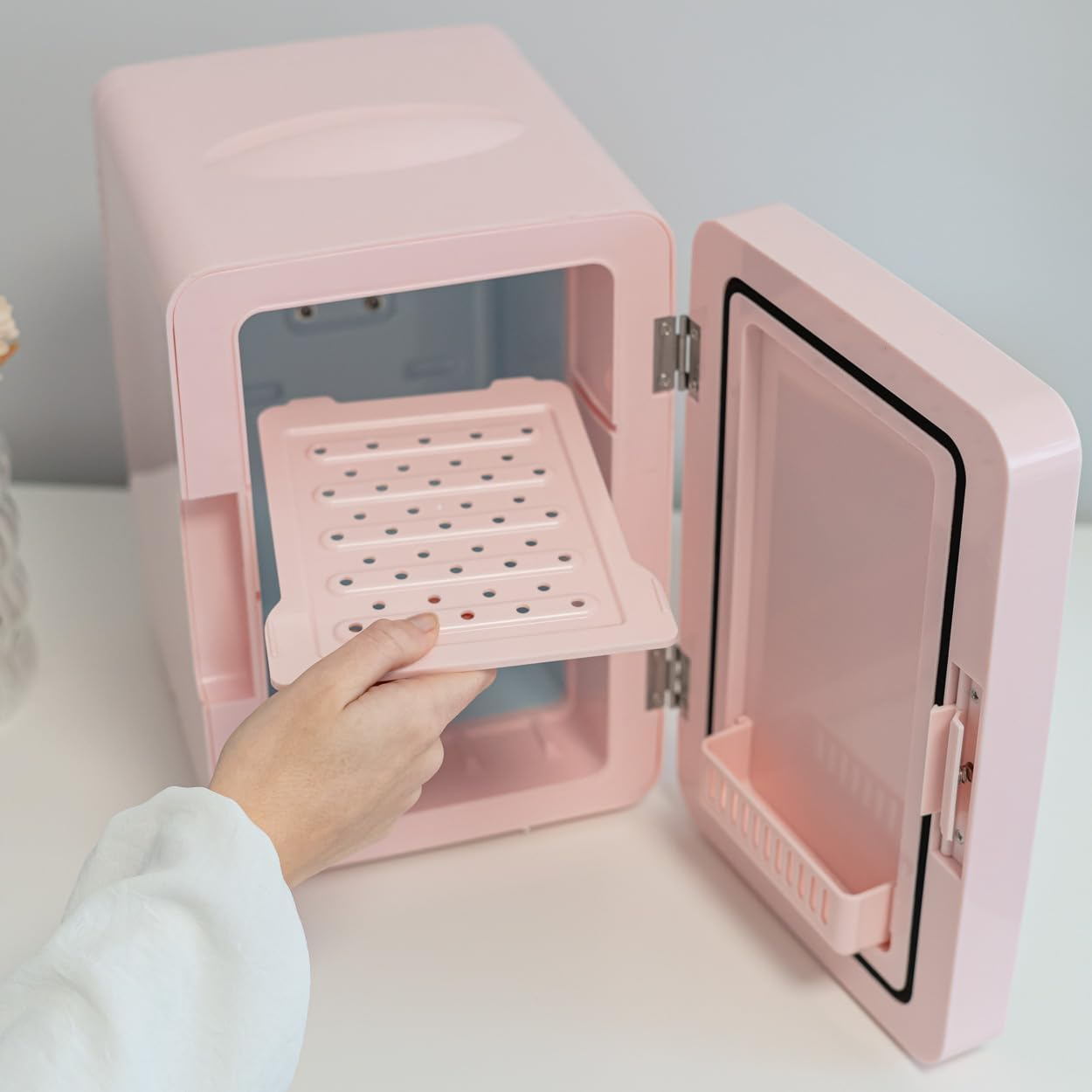 Mini nevera rosa con la puerta abierta, una mano retira una bandeja. Superficie blanca.