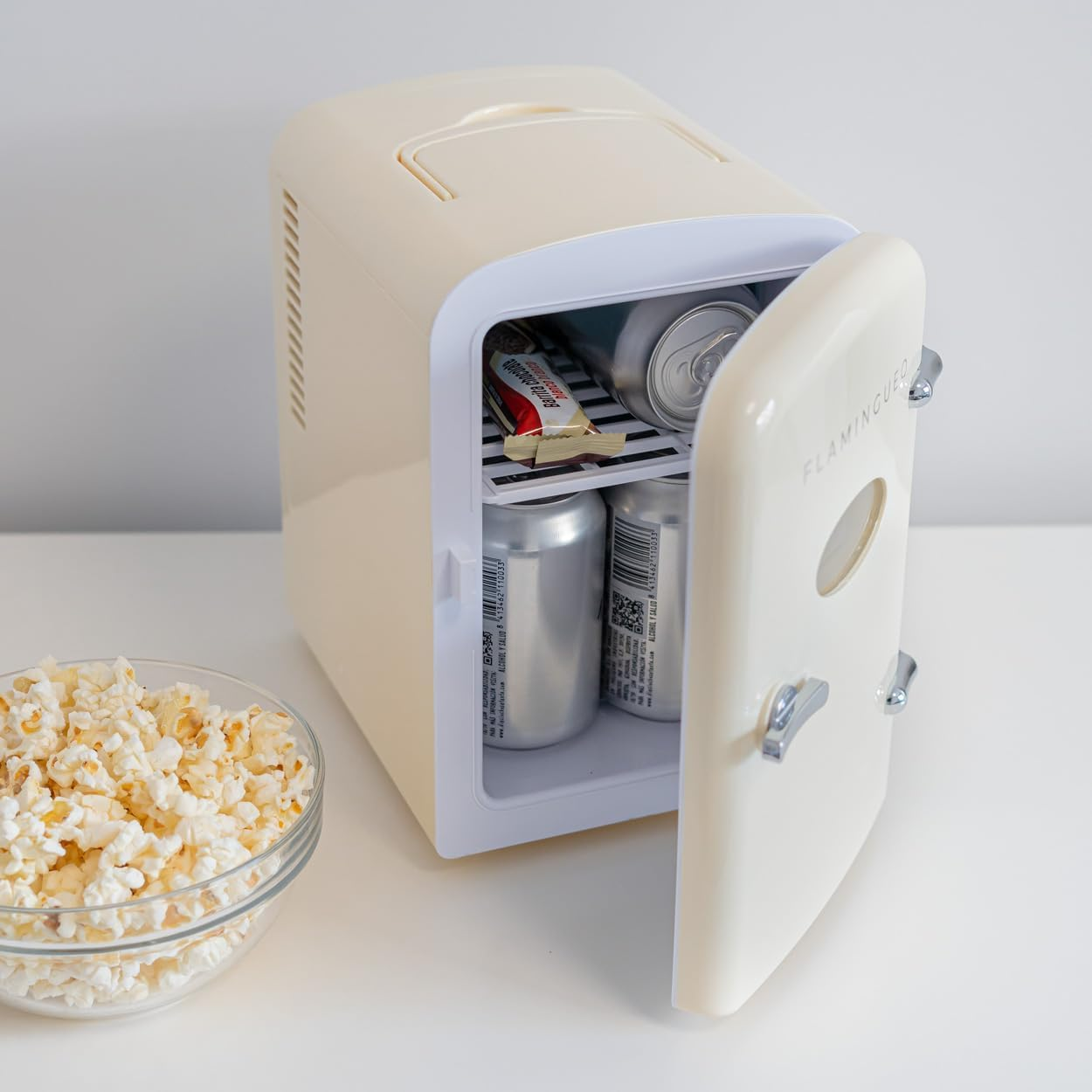 Una mini-nevera crema con la puerta abierta, con bebidas y un aperitivo. Un cuenco de palomitas en primer plano.