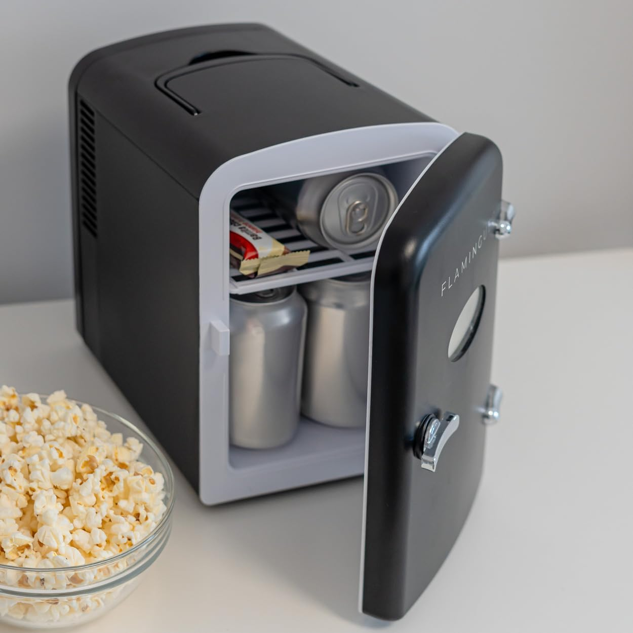 Mini nevera en blanco y negro con puerta abierta, mostrando bebidas y un snack.
