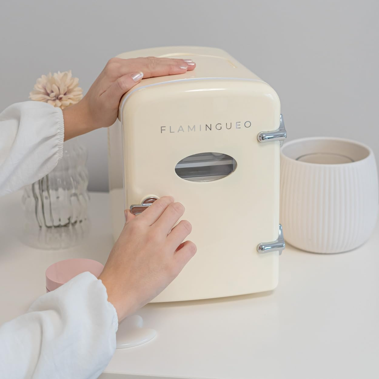 Una persona abre una mini nevera crema sobre una mesa blanca con una flor y un jarrón.