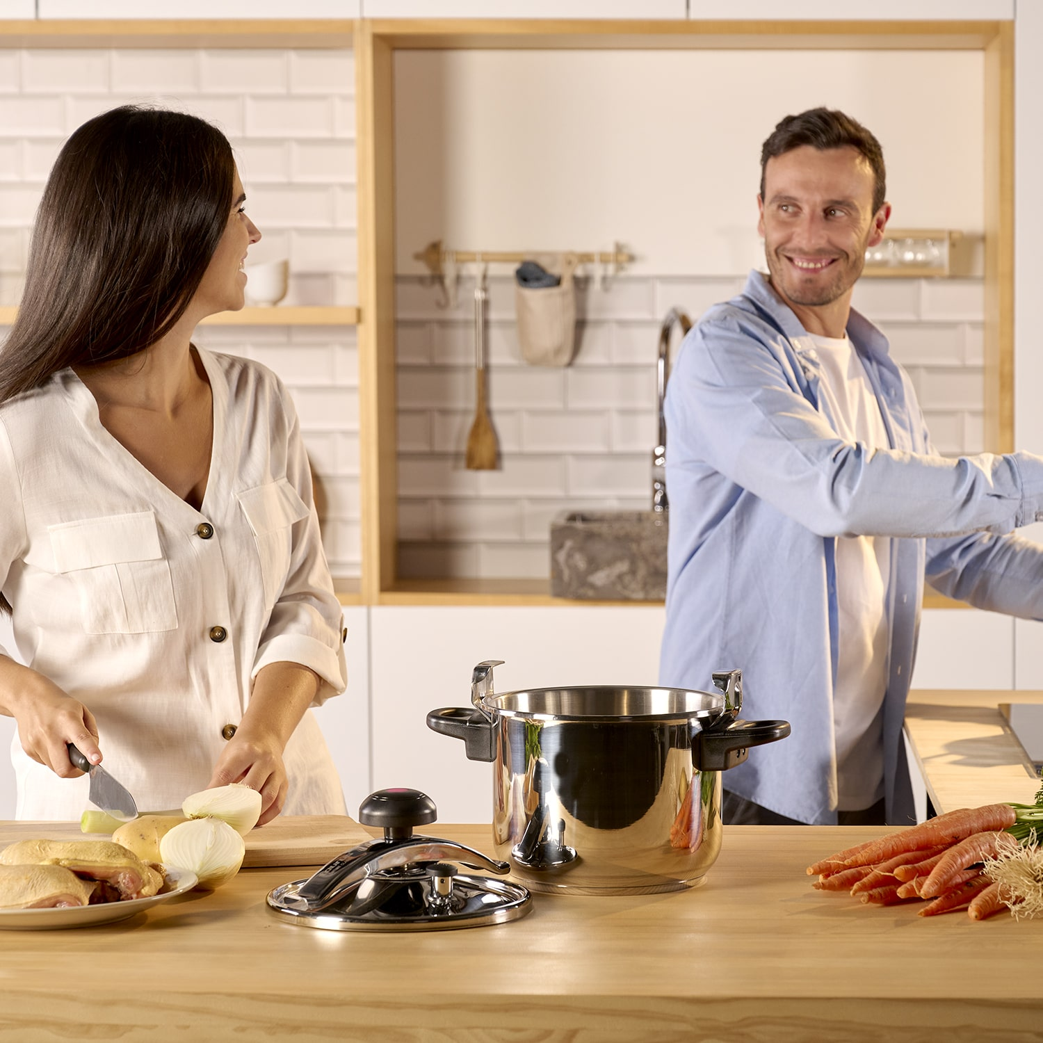 Una pareja cocinando junta. Una mujer cortando cebollas, un hombre mirando una olla. Comida en la mesa.