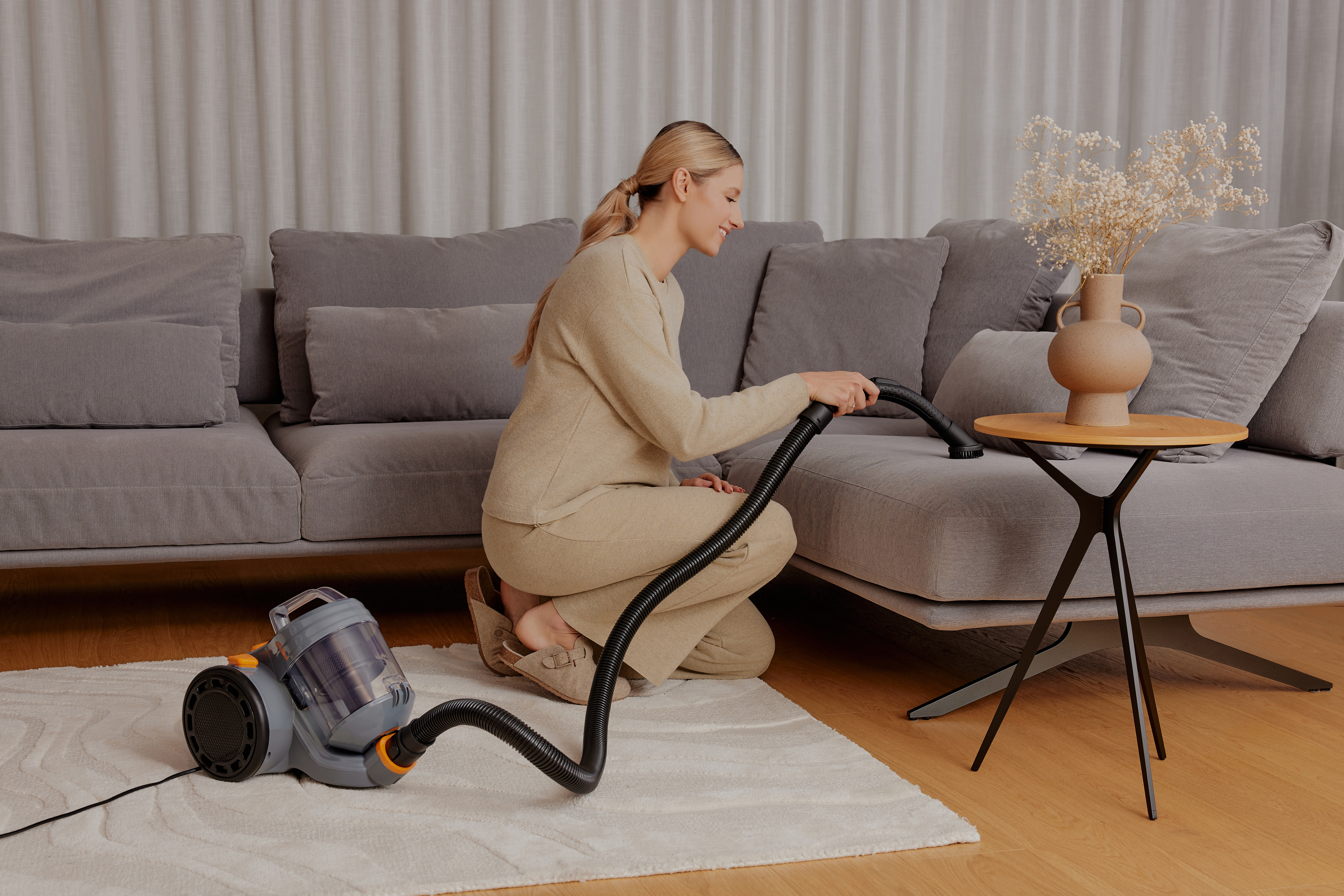 Frau, die einen grauen Sofa absaugt, mit einer Vase Blumen auf einem Tisch.
