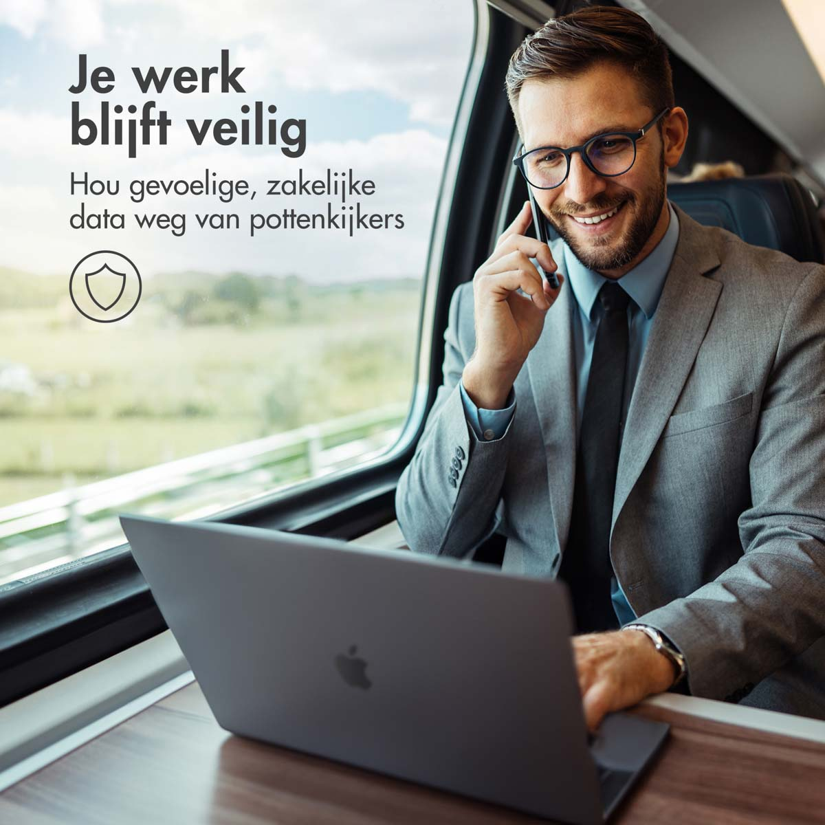 Man in a suit on a train, using a laptop and phone. Window and trees are in the background.