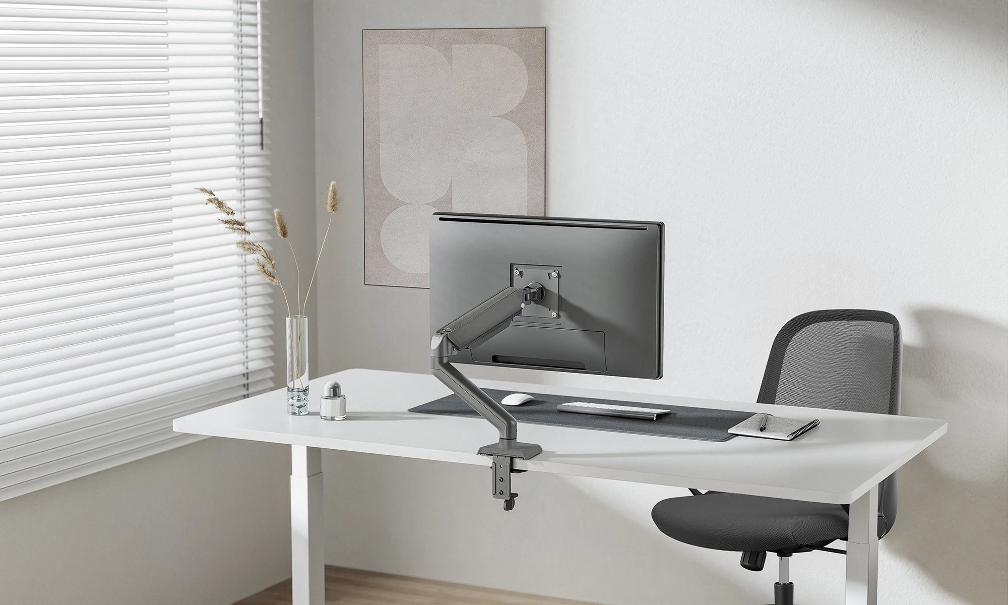 Schreibtisch-Setup: Monitor, Monitorarm, Maus, Tastatur, Notizbuch und eine Vase. Weißer Schreibtisch, grauer Stuhl.