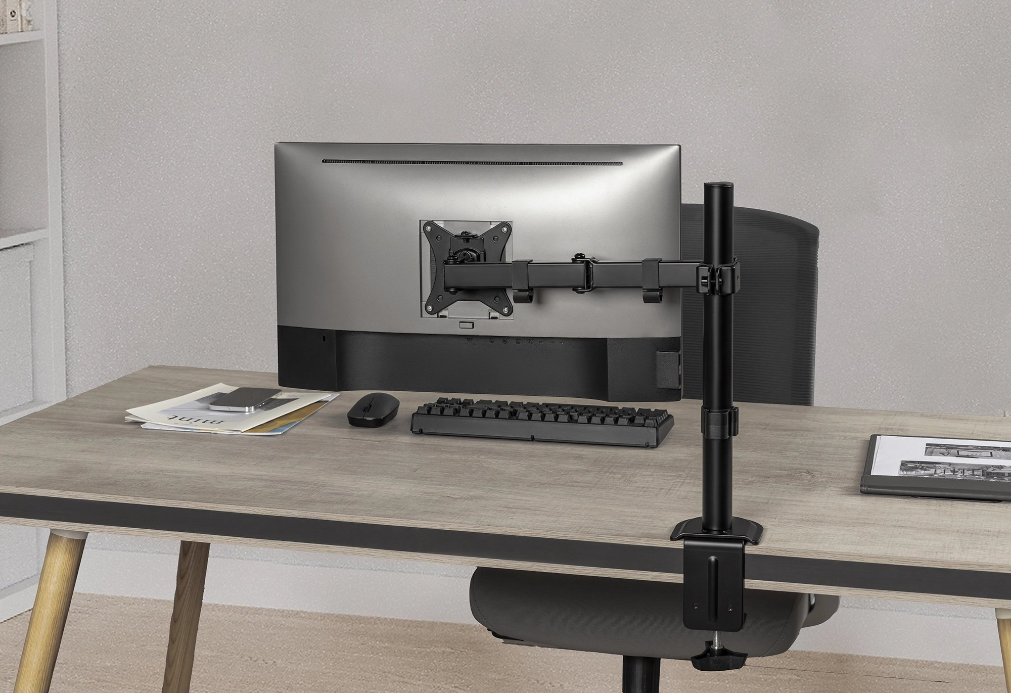 Ein Computermonitor, der mit einem schwarzen Arm an einem Schreibtisch befestigt ist. Tastatur und Maus sichtbar.