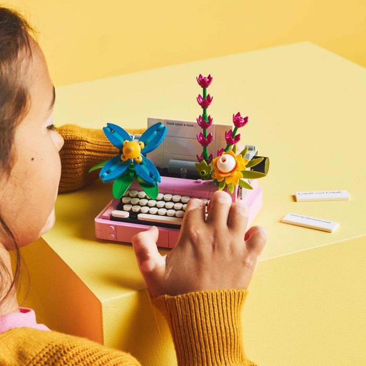 Un niño juega con un modelo de máquina de escribir Lego con flores. Fondo amarillo con bloques de texto.