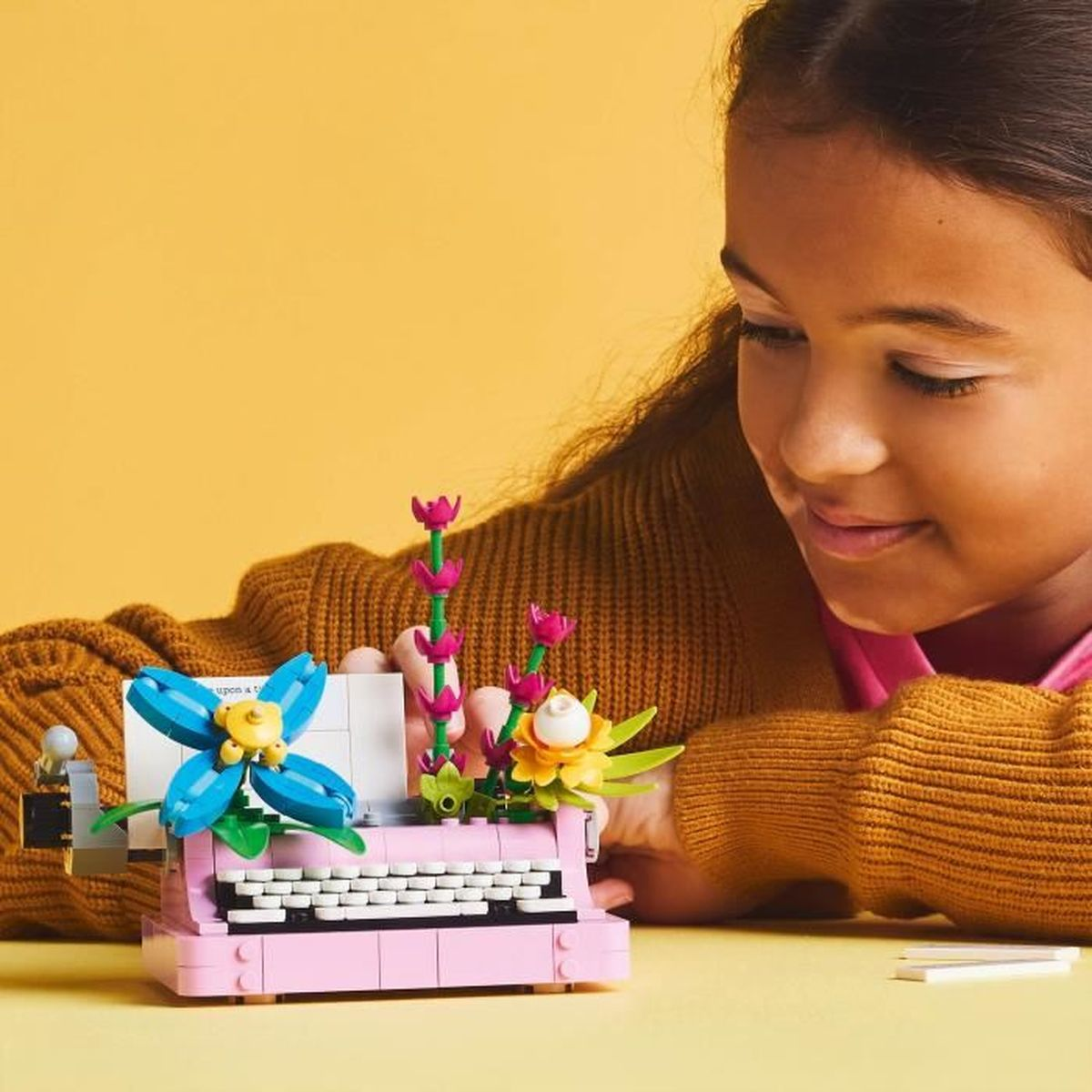 Niña con máquina de escribir Lego con flores. Fondo amarillo.