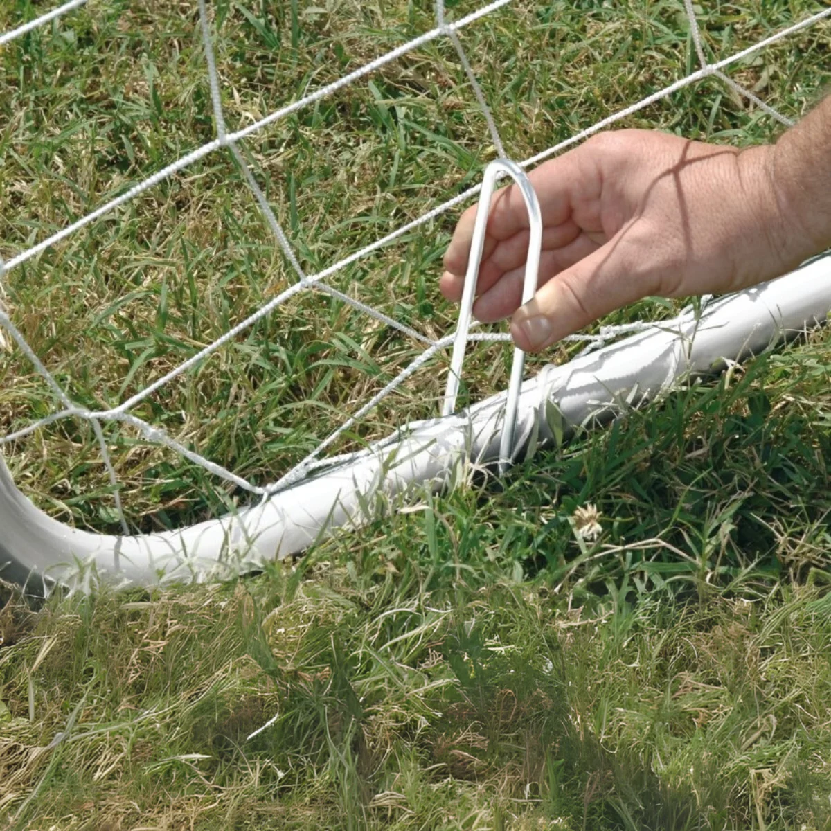 A hand is placing a metal stake into the ground to secure a soccer net, green grass in view.