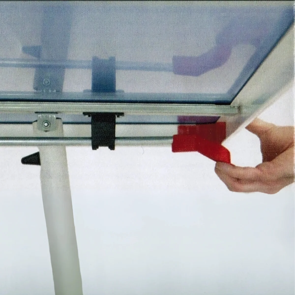 A hand pulls a red lever on a white table. A black clip is nearby. Clear panel on top.