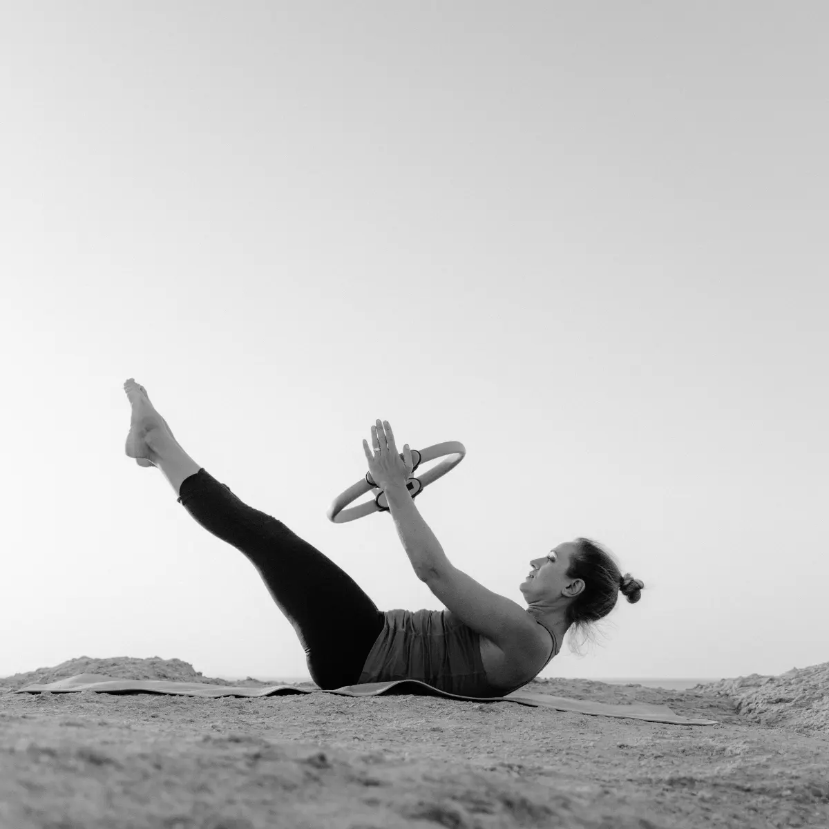 Femme faisant du Pilates sur des rochers, tenant un anneau, jambes levées. Monochrome, fond de ciel.