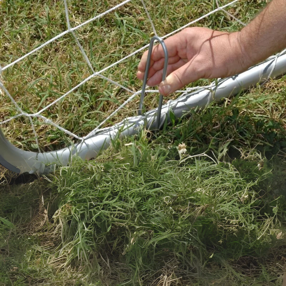 Main fixant un filet de football au sol avec un piquet métallique, gros plan.