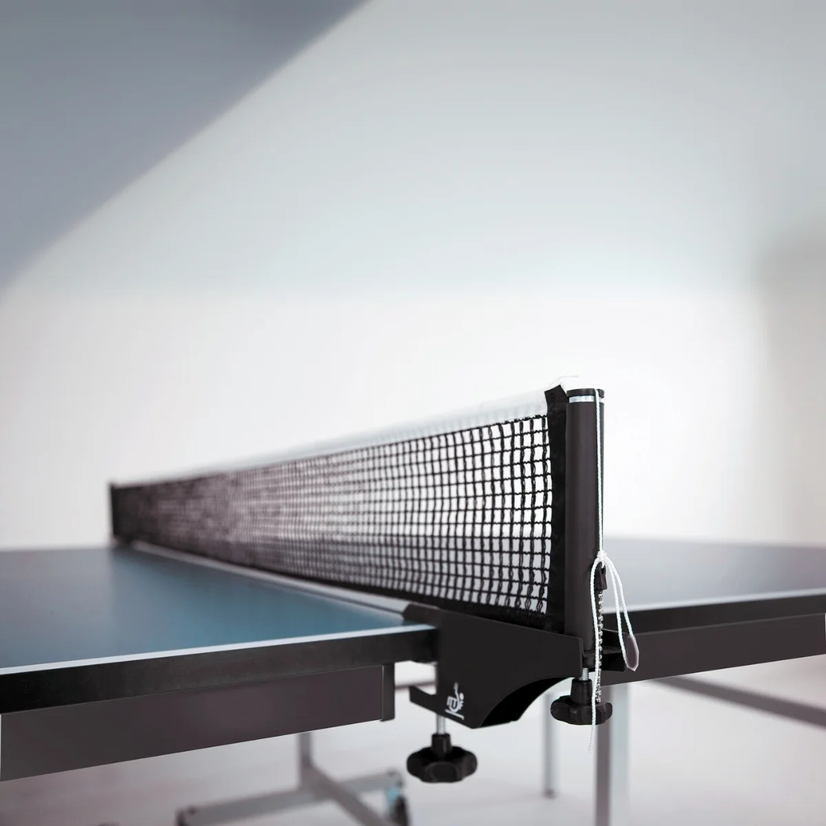 A ping pong table net. The net is black. The table is blue. White wall in the background.