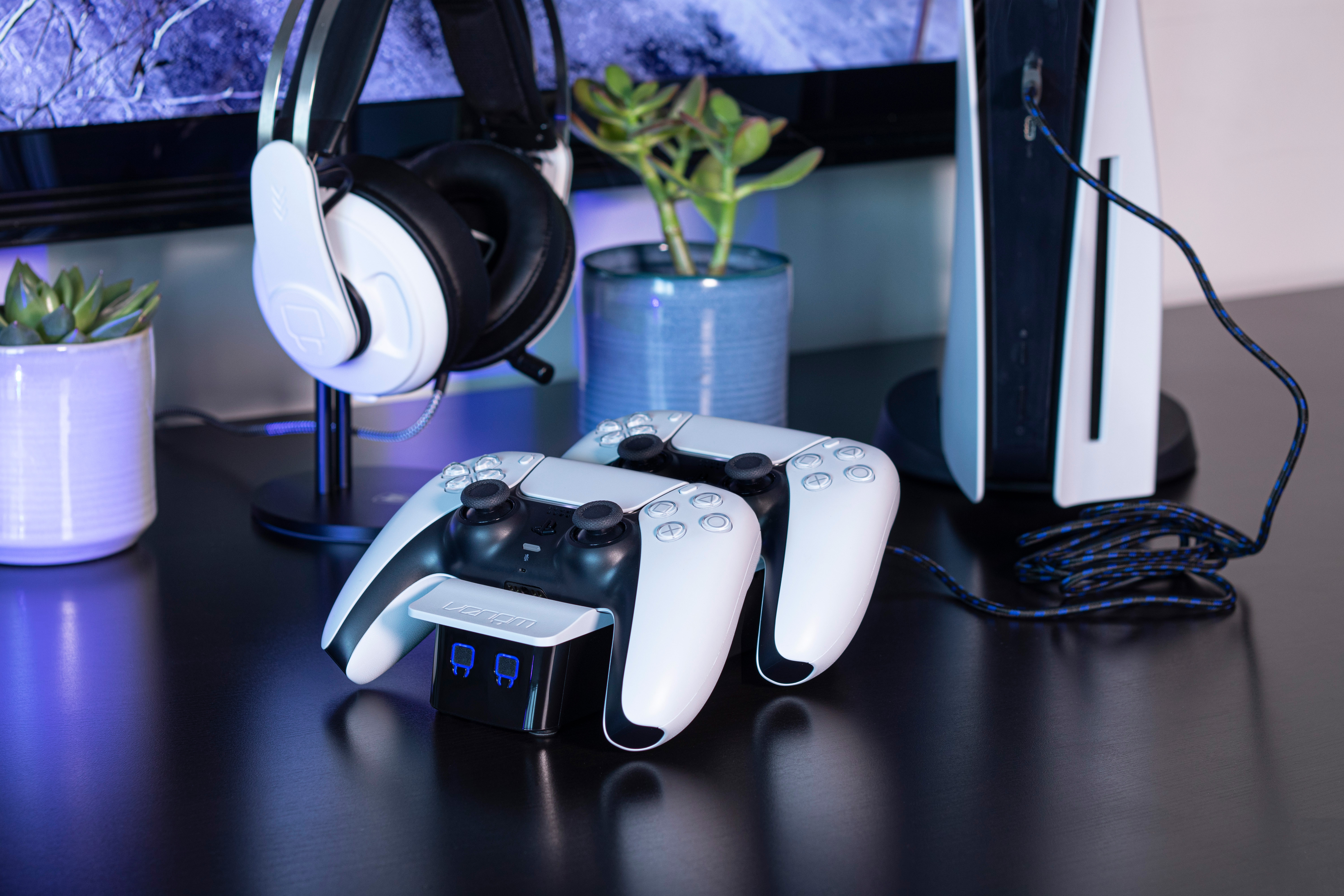 Twee witte en zwarte gamecontrollers op een oplaadstation met een headset en een plant op de achtergrond.