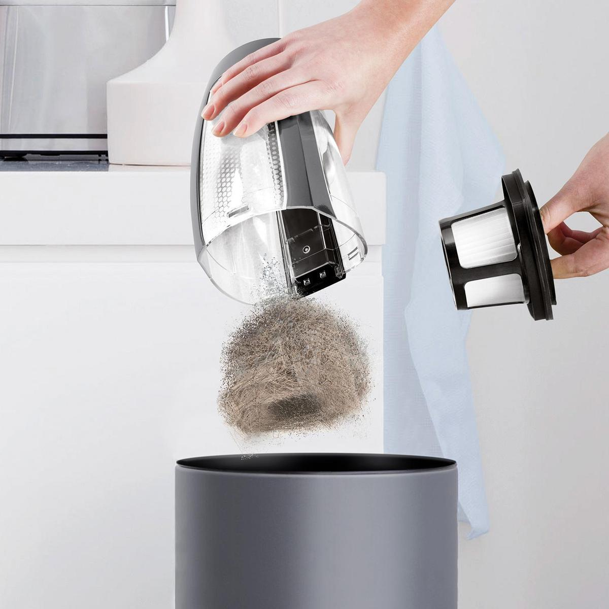 Hände leeren einen Staubsaugerbehälter in einen Mülleimer. Behälter und Filter sind sichtbar.