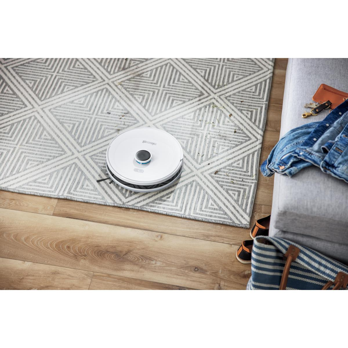 Un aspirateur robot blanc sur un tapis à motifs, à côté de meubles et d'autres objets.