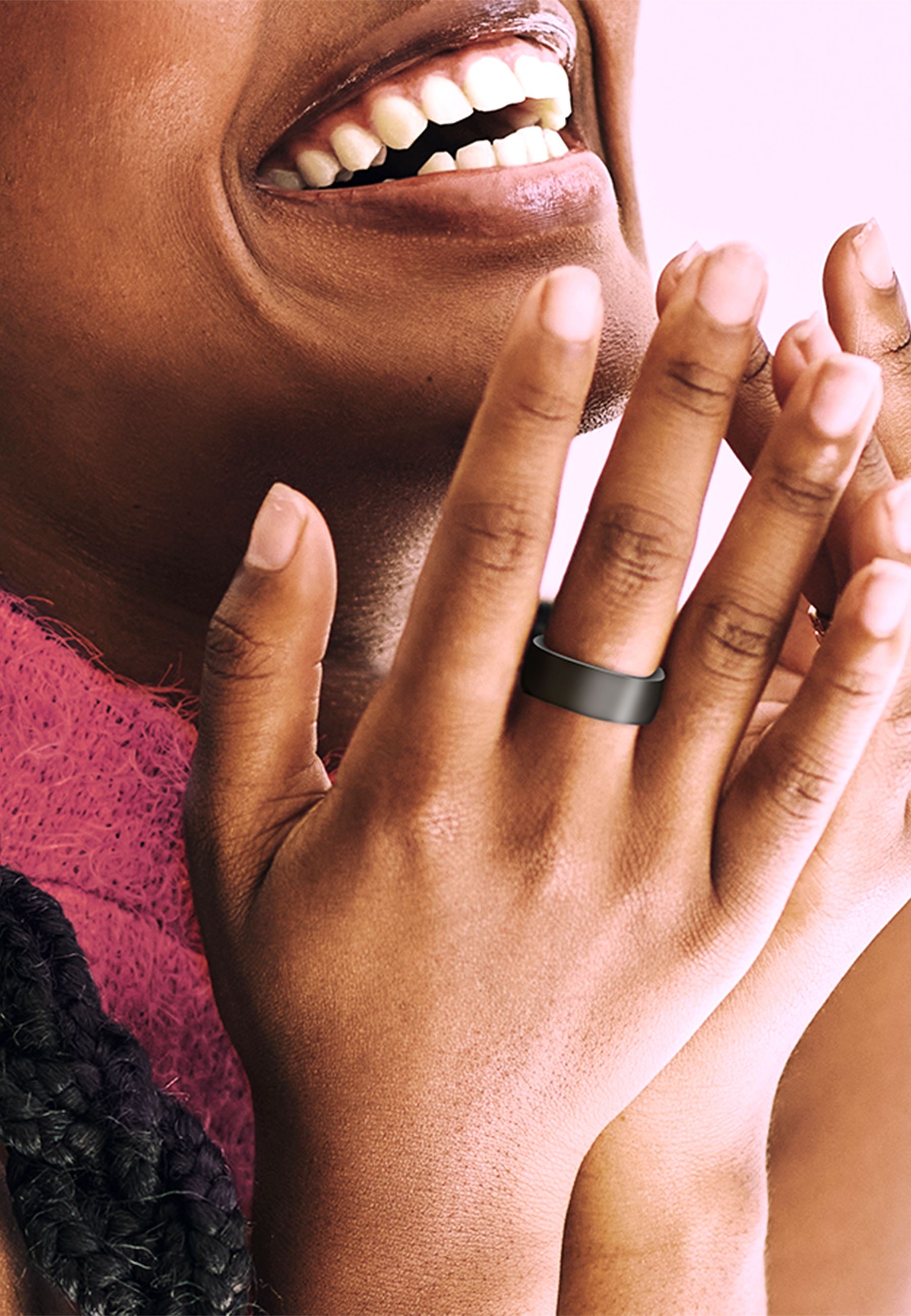 Die Hand einer Person mit einem schwarzen Ring am Finger, lächelnd, vor rosa Hintergrund.
