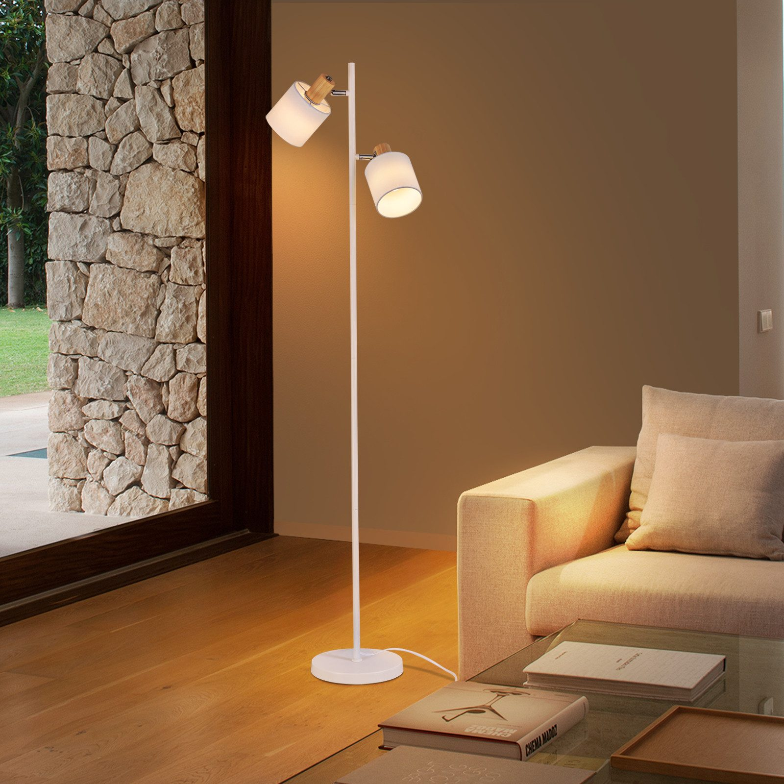 Eine Lampe mit zwei Lichtern an einem Ständer. Sie steht neben einem Fenster und einem beigefarbenen Sofa.