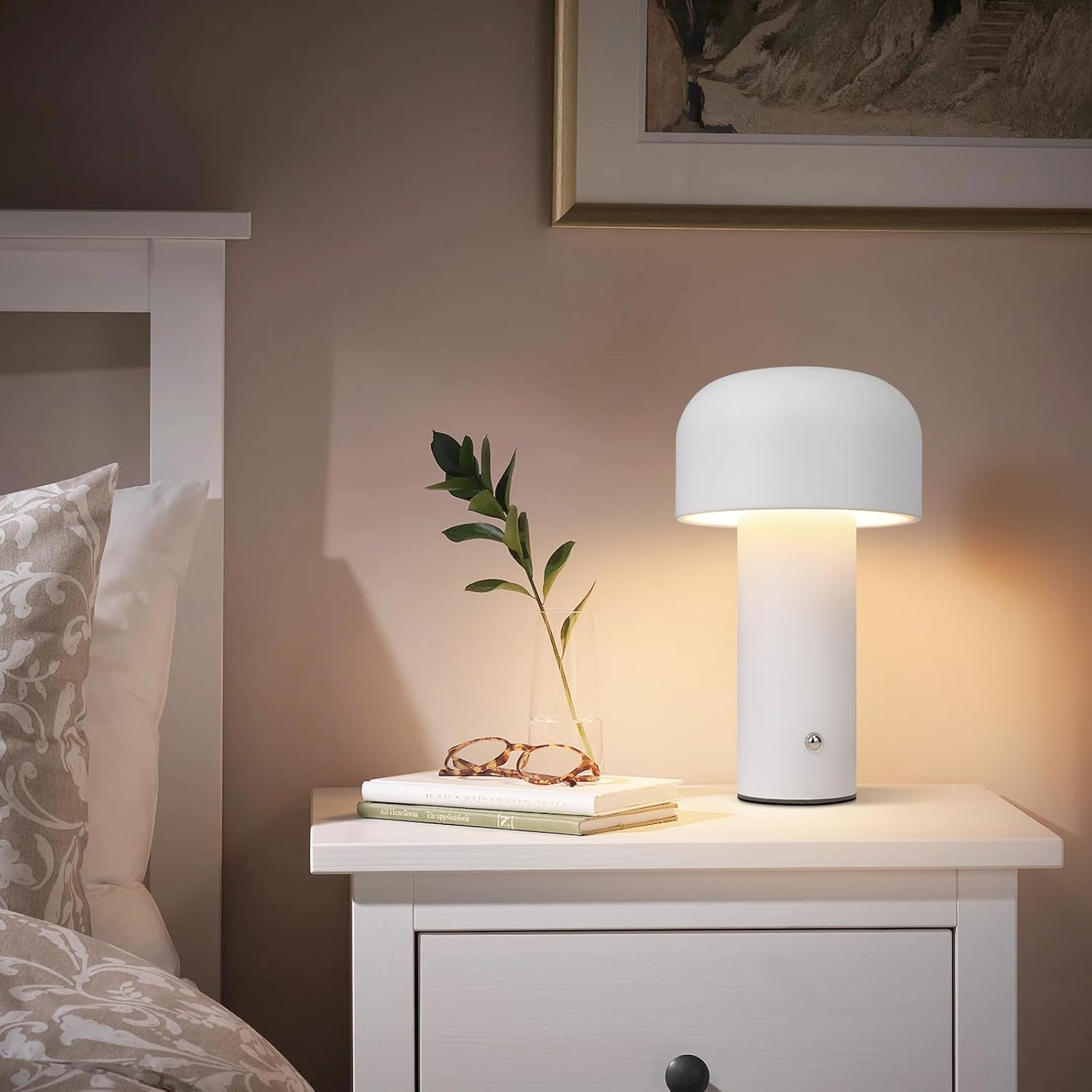 Biała lampka nocna na stoliku nocnym. Jest szklany wazon i książka. Łóżko jest w tle.