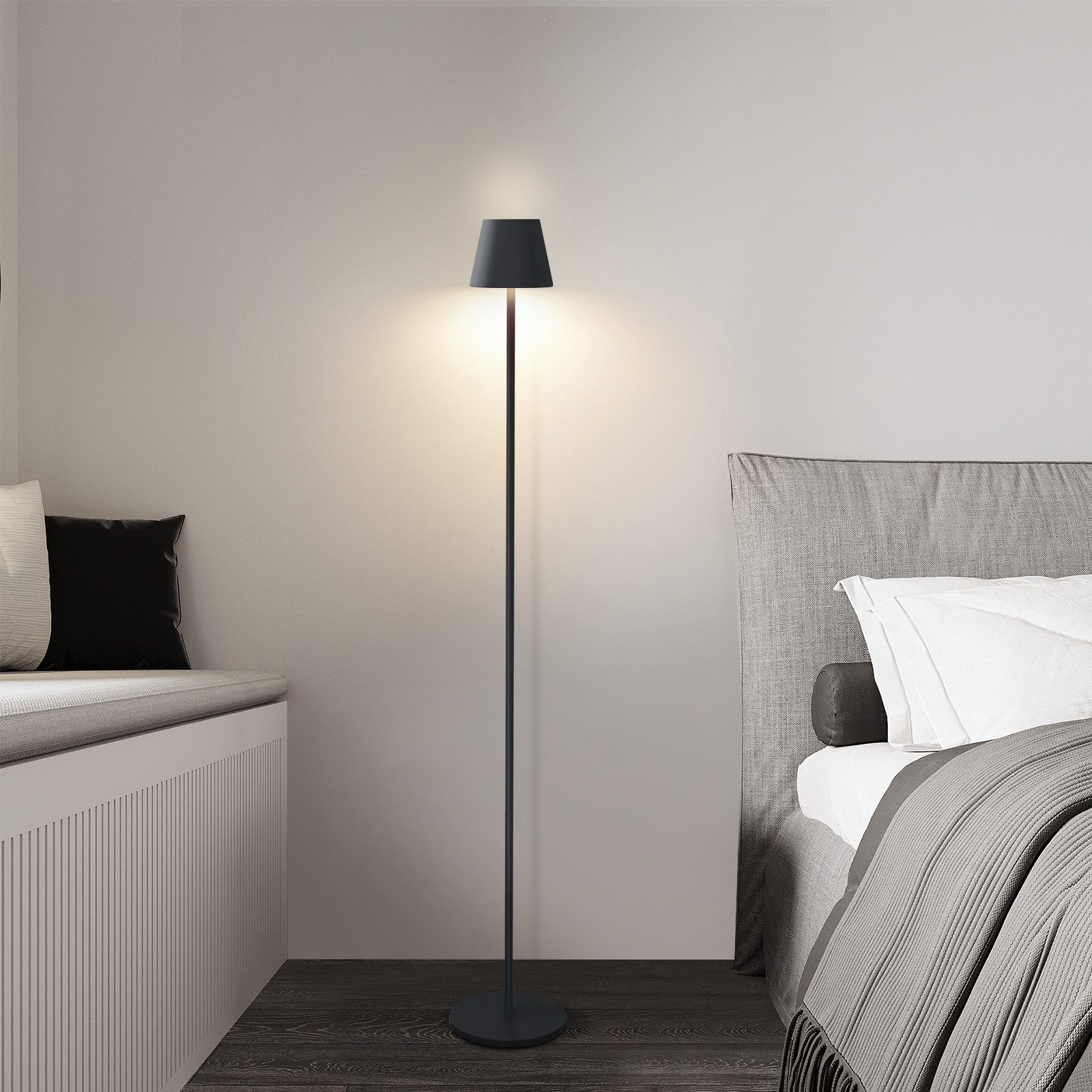Una lámpara de pie negra con pantalla, de pie cerca de una cama y un asiento junto a la ventana, sobre un piso de madera.