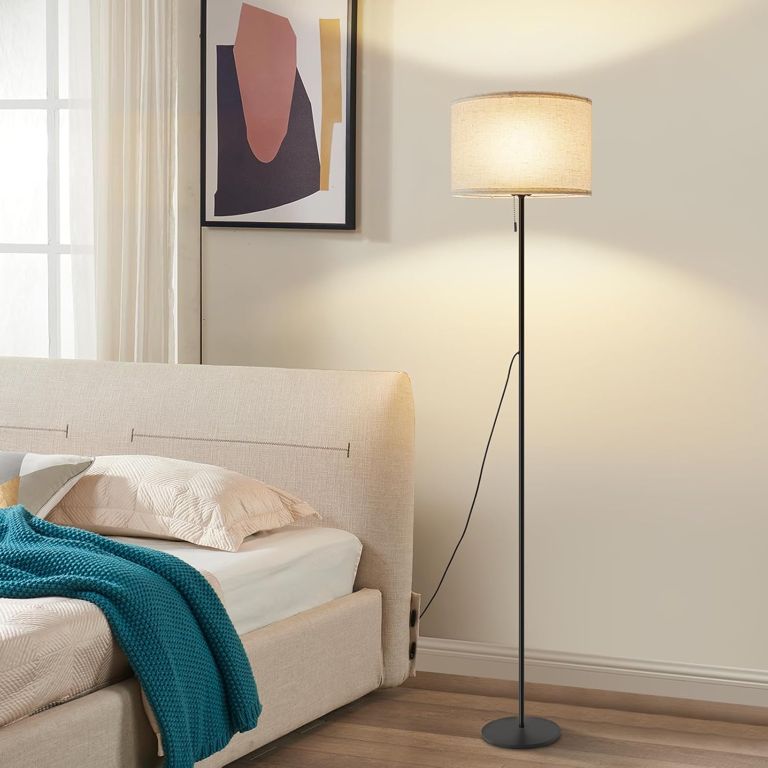 Lampa podłogowa obok łóżka. Lampa ma beżowy klosz. Obecny jest obraz i niebieski koc.