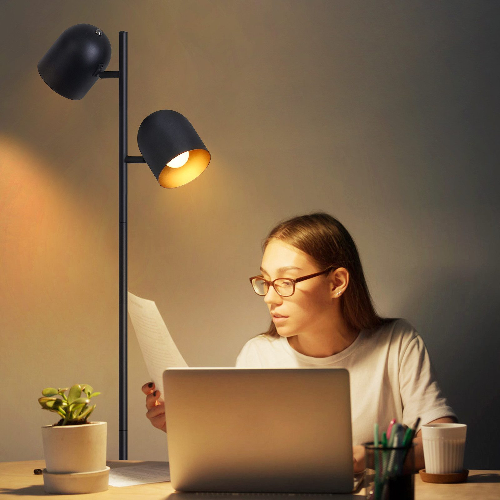 Una donna legge a una scrivania con un laptop e una lampada da terra con due paralumi neri.