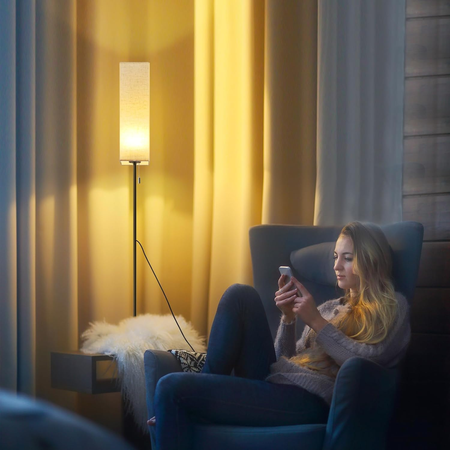 Kobieta w fotelu używająca telefonu, z lampą i zasłonami w tle.